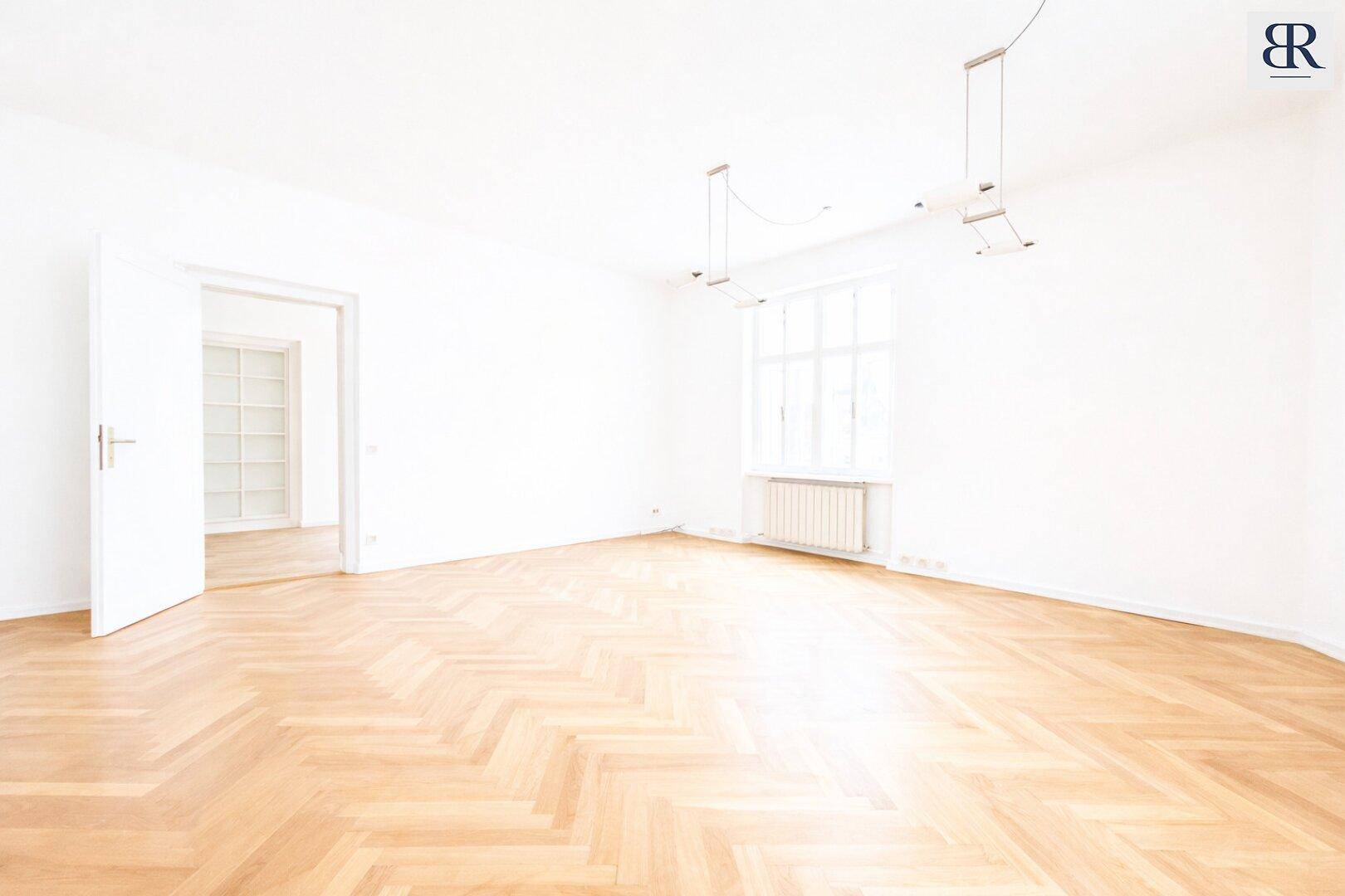 Geräumiger, heller Wohnbereich mit Fischgrätparkett und offener Tür zu einem weiteren Zimmer.