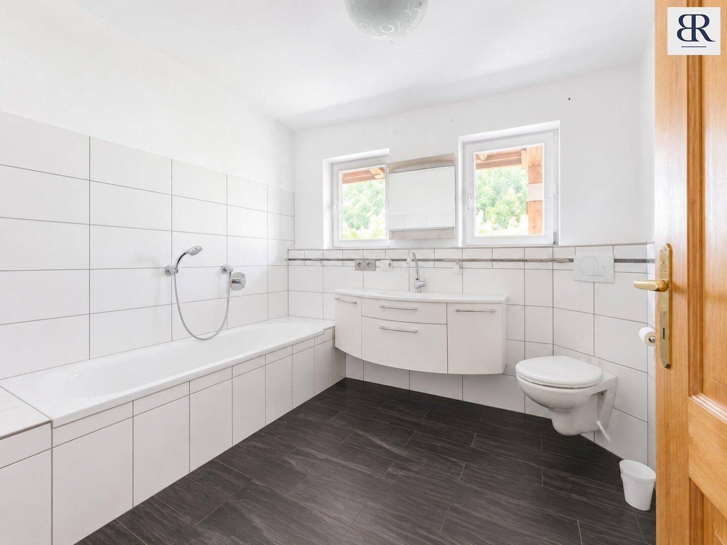 Modernes Badezimmer mit Badewanne, Waschbecken, WC und dunklen Bodenfliesen, gut beleuchtet.