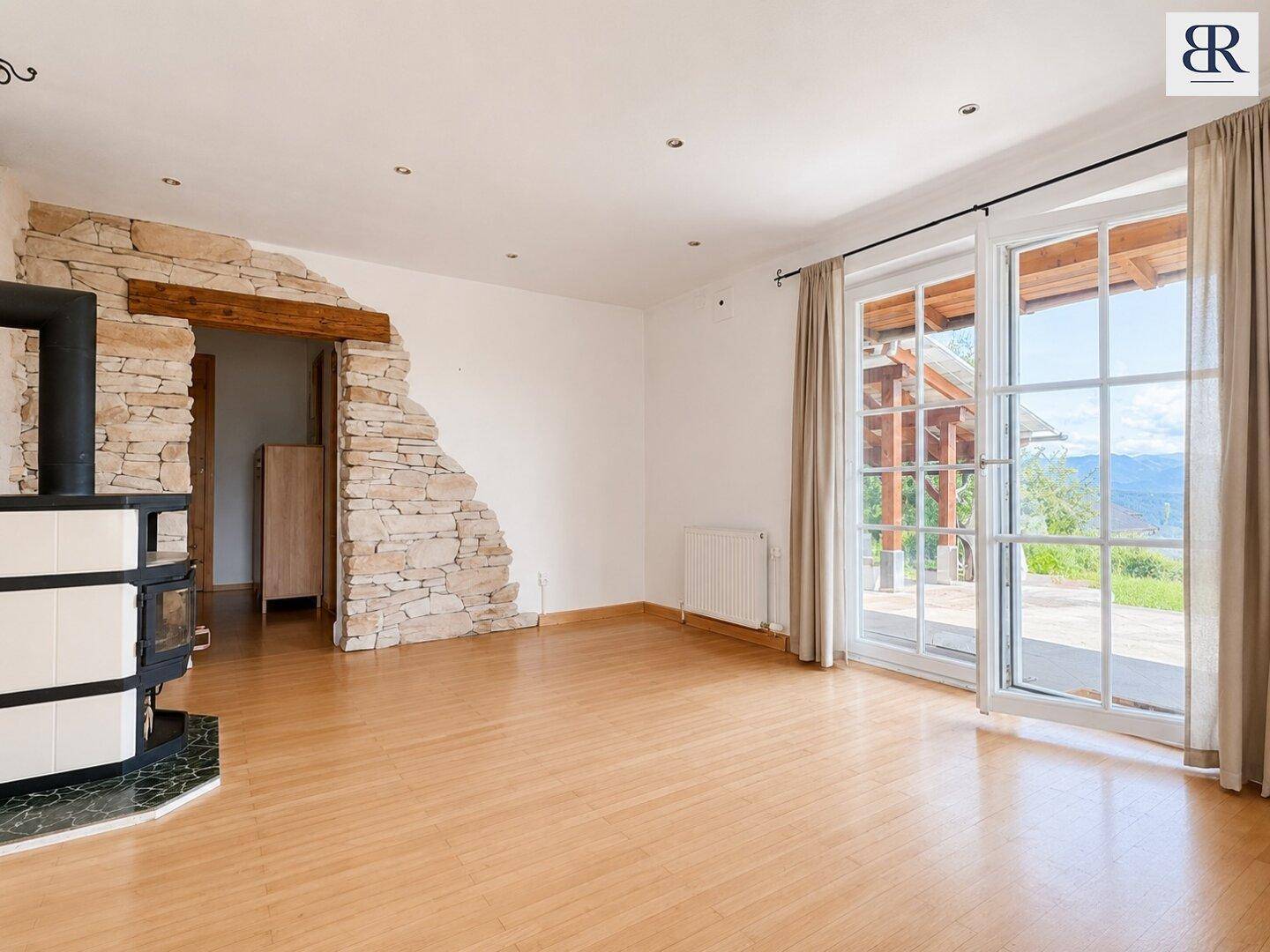 Helles Wohnzimmer mit traditionellem Kachelofen, Holzboden und Zugang zur Terrasse mit Bergblick.