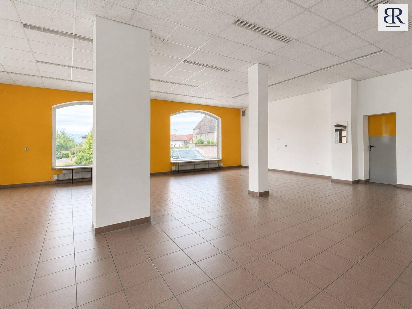 Geräumiger, offener Gewerberaum mit tragenden Säulen, großen Fenstern und Fliesenboden.