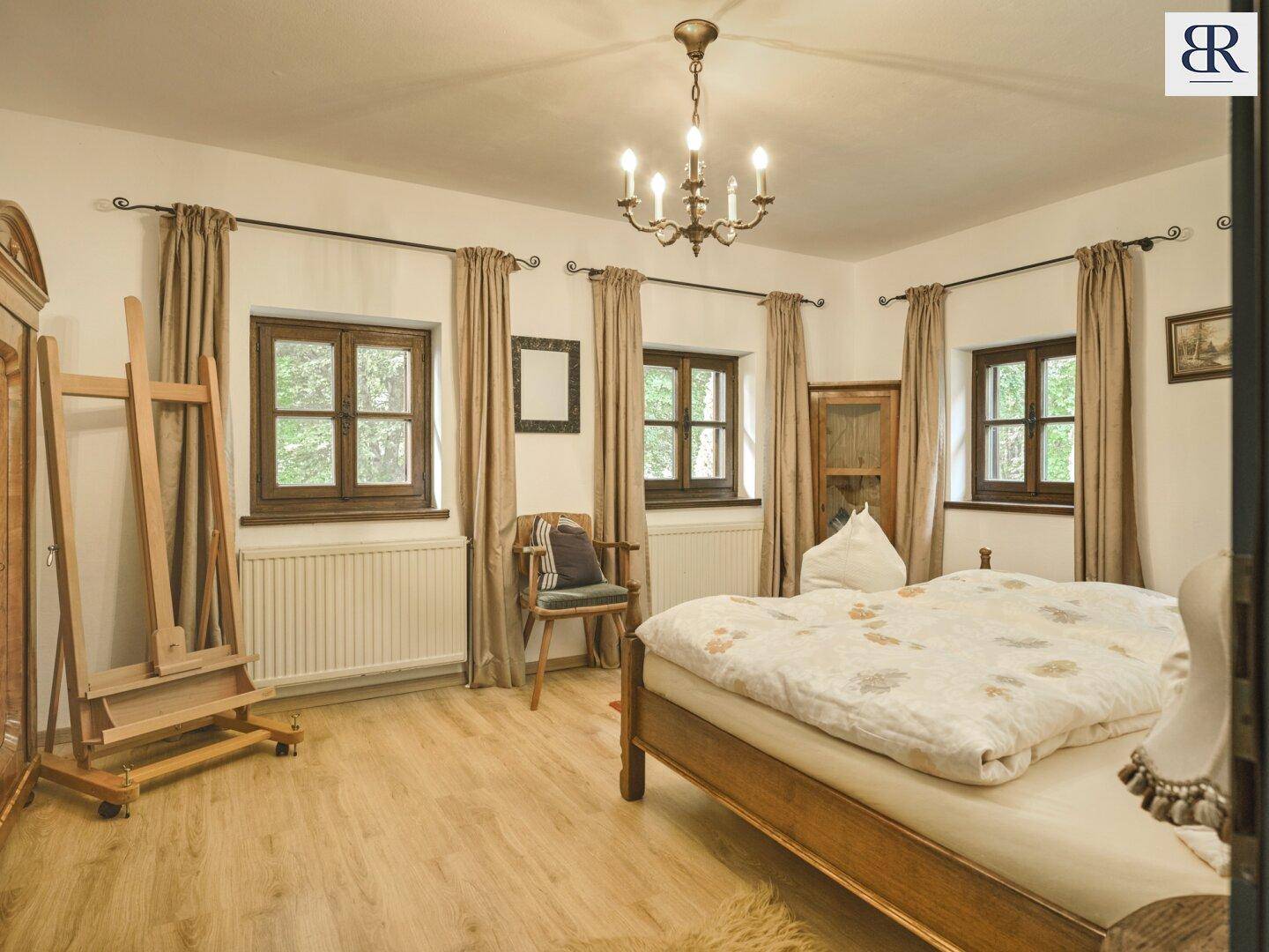 Helles Schlafzimmer mit Holzbett, hellem Holzboden und mehreren Fenstern mit Blick ins Grüne.