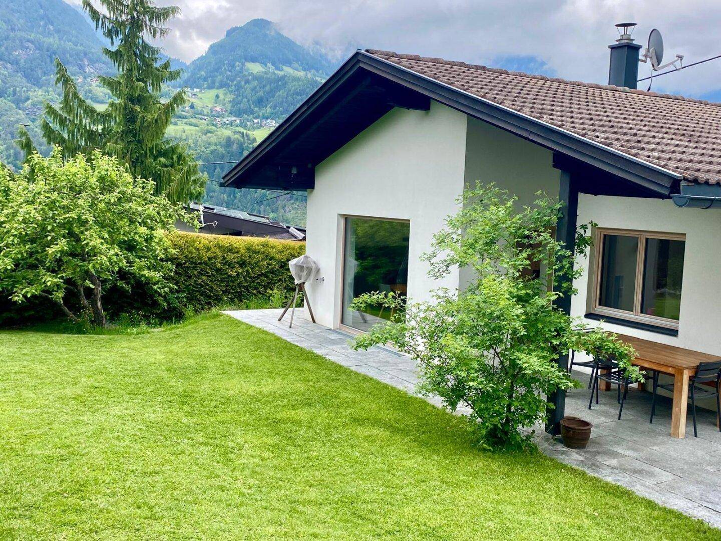 Gepflegter Gartenbereich mit Terrasse und Sitzgelegenheiten, umgeben von grüner Natur und Bergen.