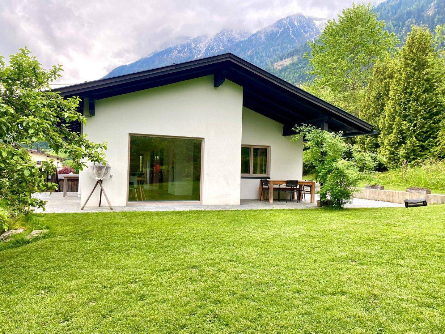Modernes Haus mit großem Garten und Terrasse, umgeben von einer beeindruckenden Berglandschaft.