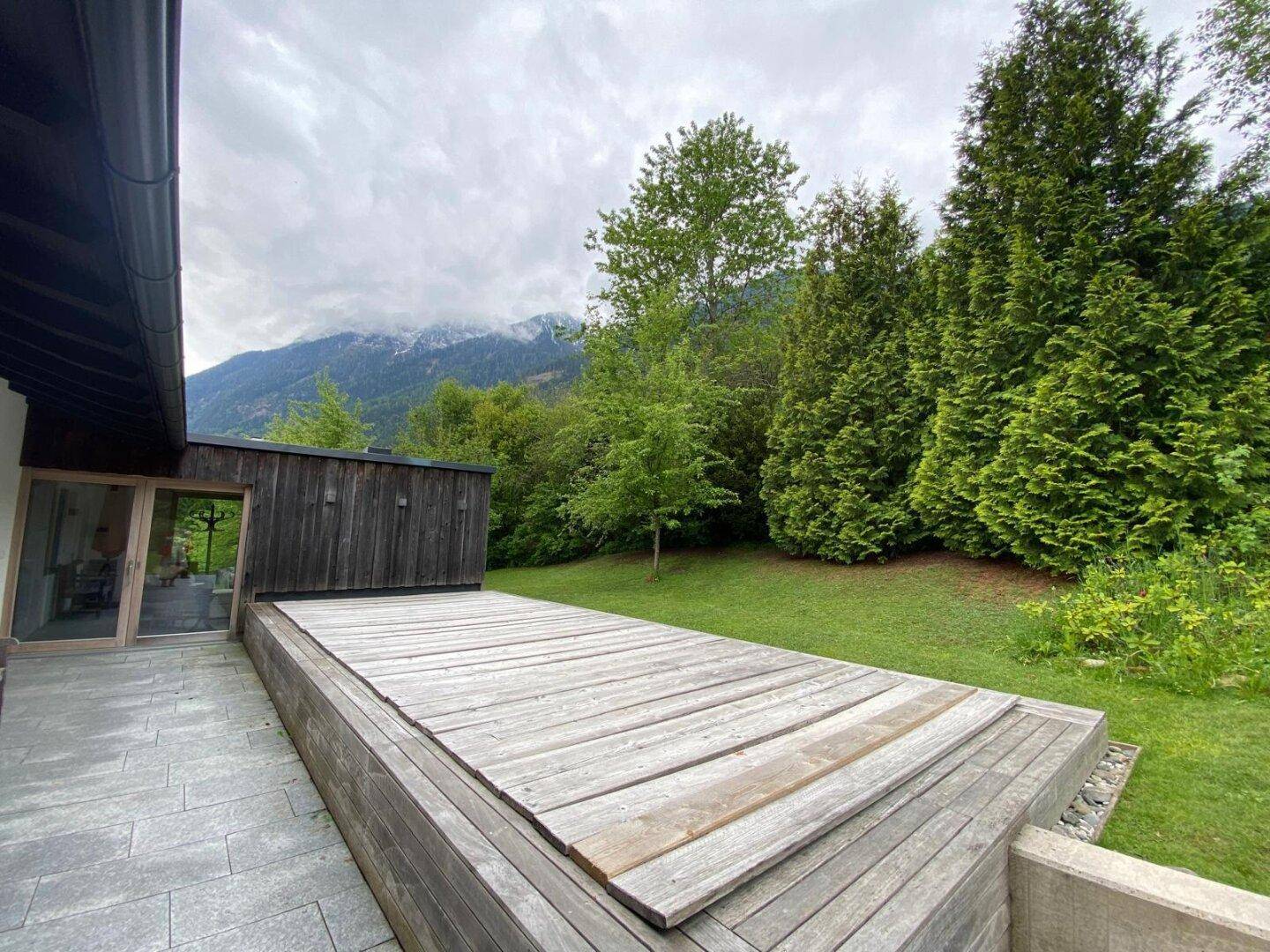 Holzterrasse mit Blick auf den Garten und die bewaldeten Hänge der Berge.