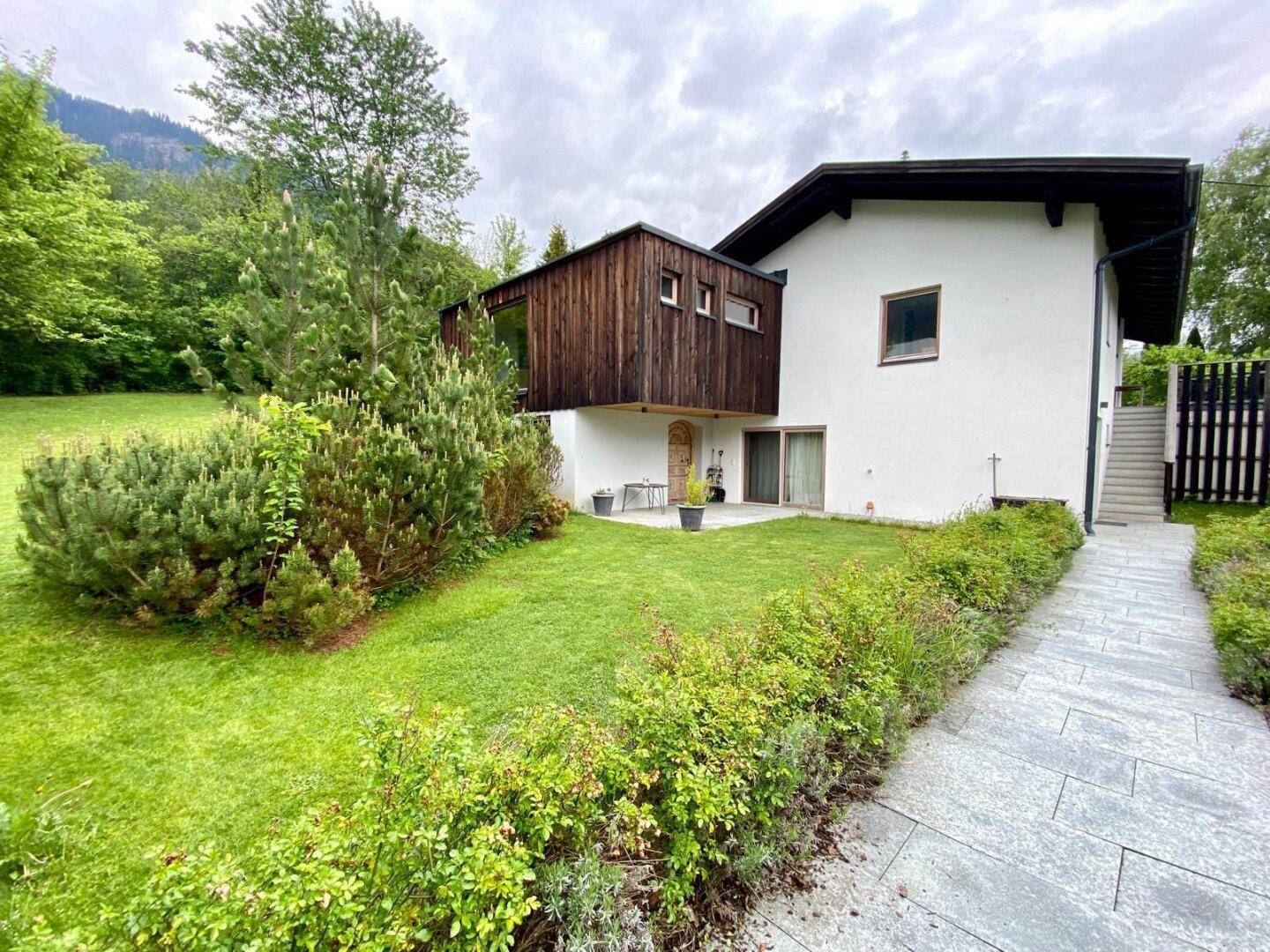 Hausfassade mit gepflegtem Garten, kleiner Terrasse und Bergblick unter bewölktem Himmel.