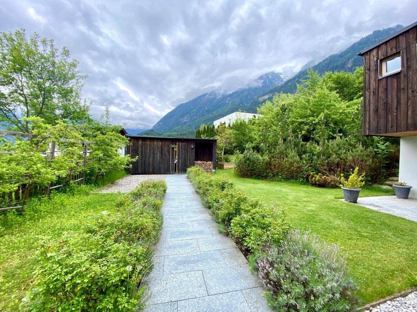 Langer Steinweg führt durch den grünen Garten zu einem Holzschuppen mit Bergblick.