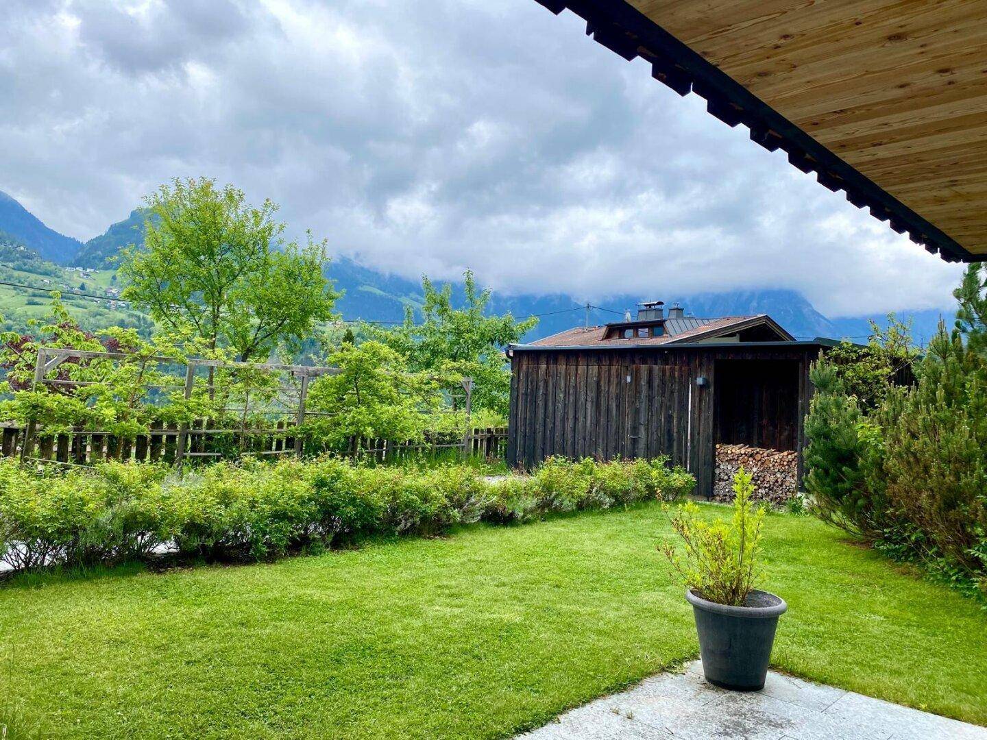 Gepflegter Garten mit Rasenfläche, Holzschuppen und beeindruckendem Bergblick unter bewölktem Himmel.