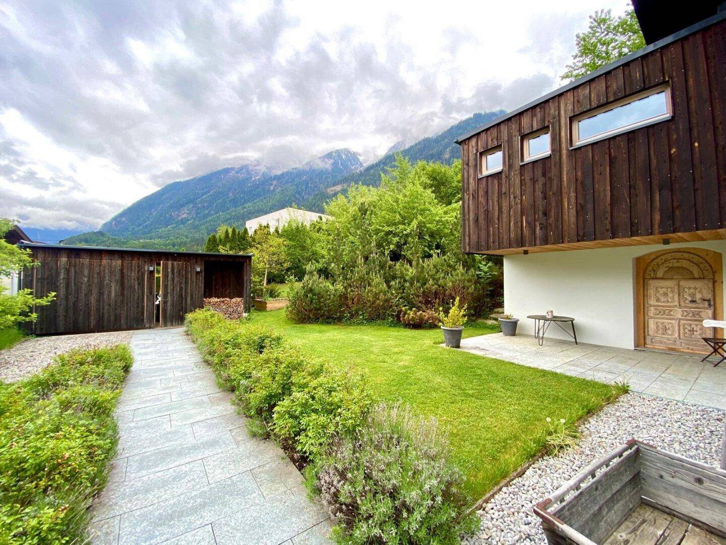 Haus mit Holzfassade und weißem Putz, gepflegtem Garten, Terrasse und Bergblick.