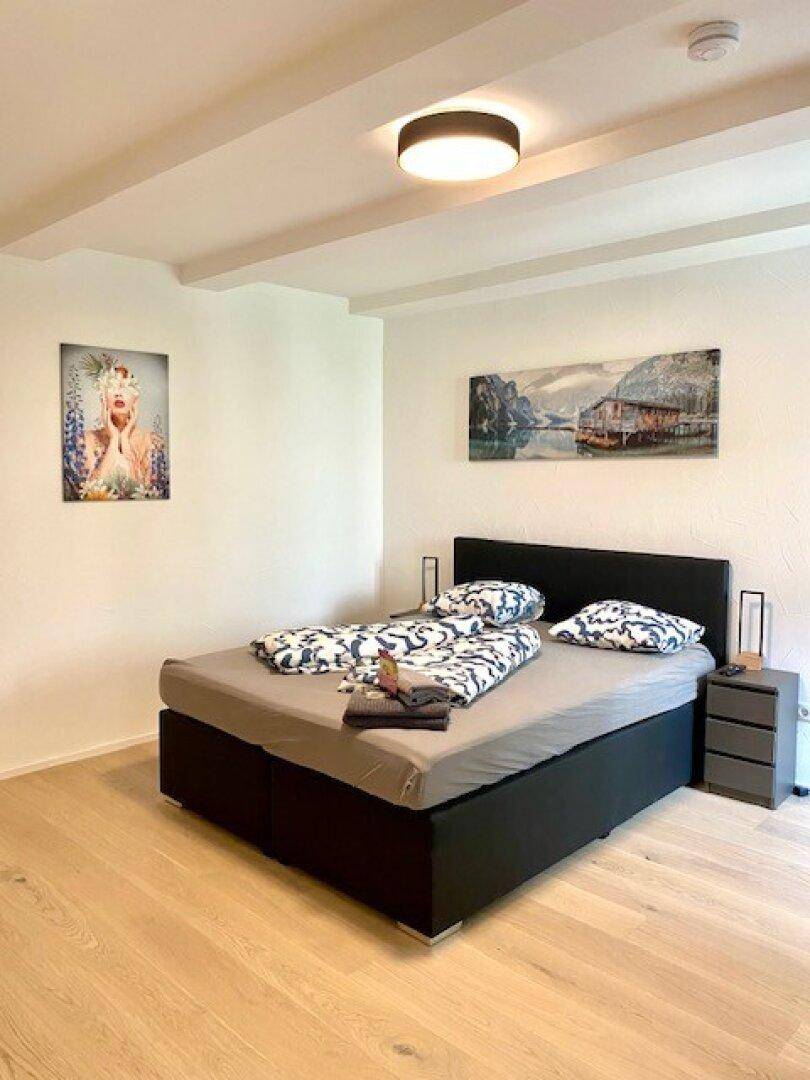 Helles Schlafzimmer mit Doppelbett, Holzboden und Kunst an den Wänden, unter sichtbaren Deckenbalken.