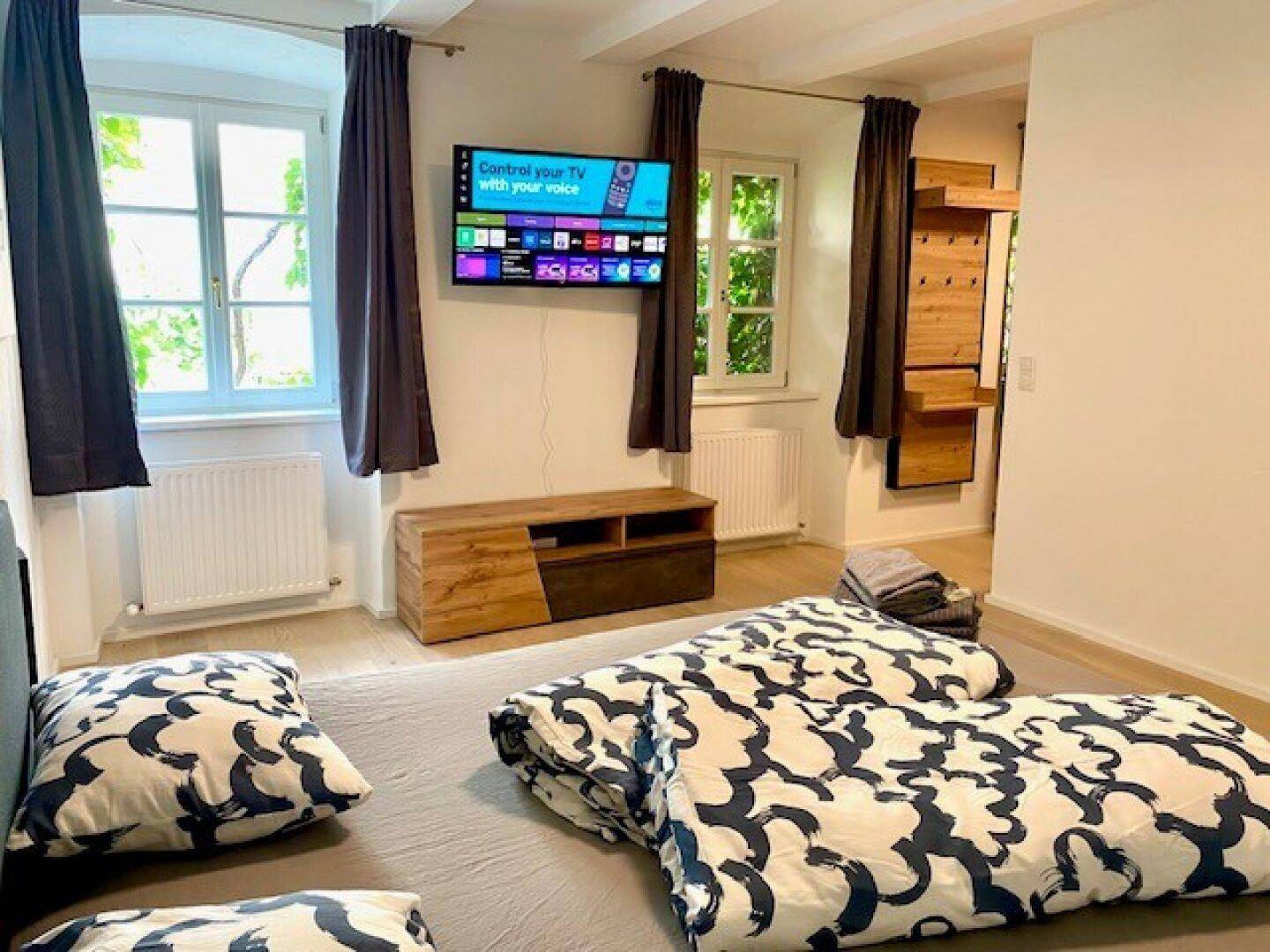 Helles Schlafzimmer mit Bodenbett, Fernseher und Holzmöbeln, durch Fenster fällt viel Tageslicht herein.