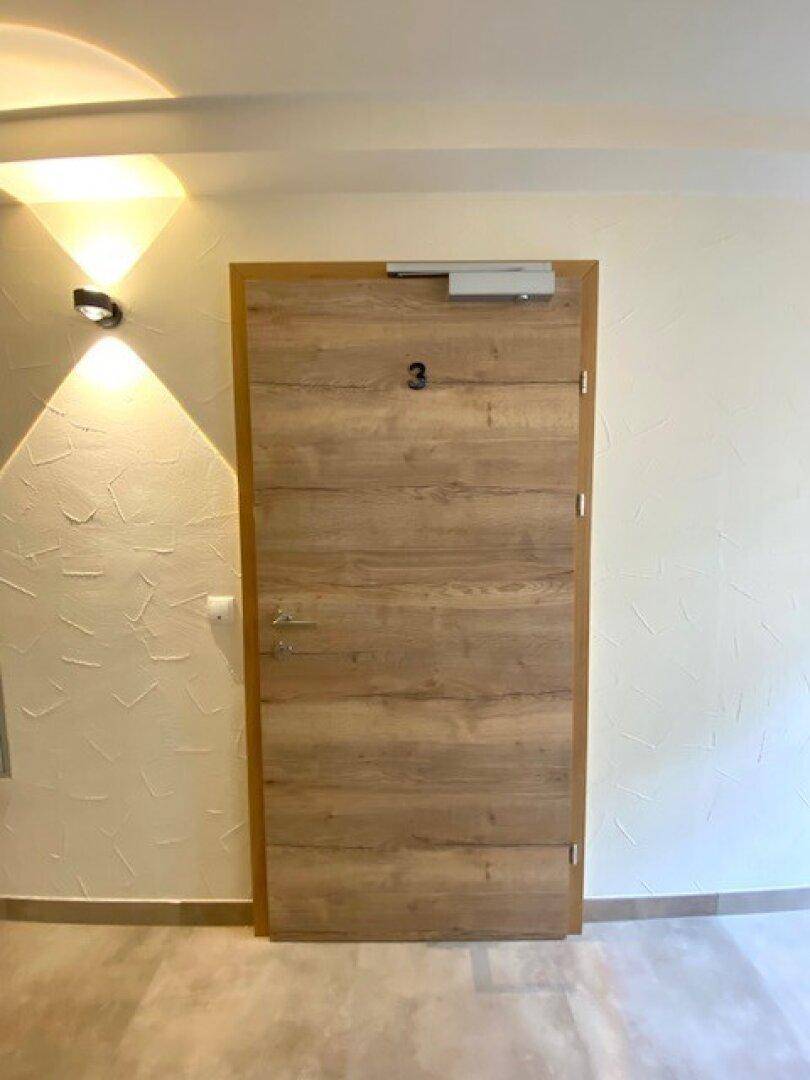 Eingangstür aus Holz mit der Nummer drei in einem hellen Flur mit Wandleuchten und Fliesenboden.
