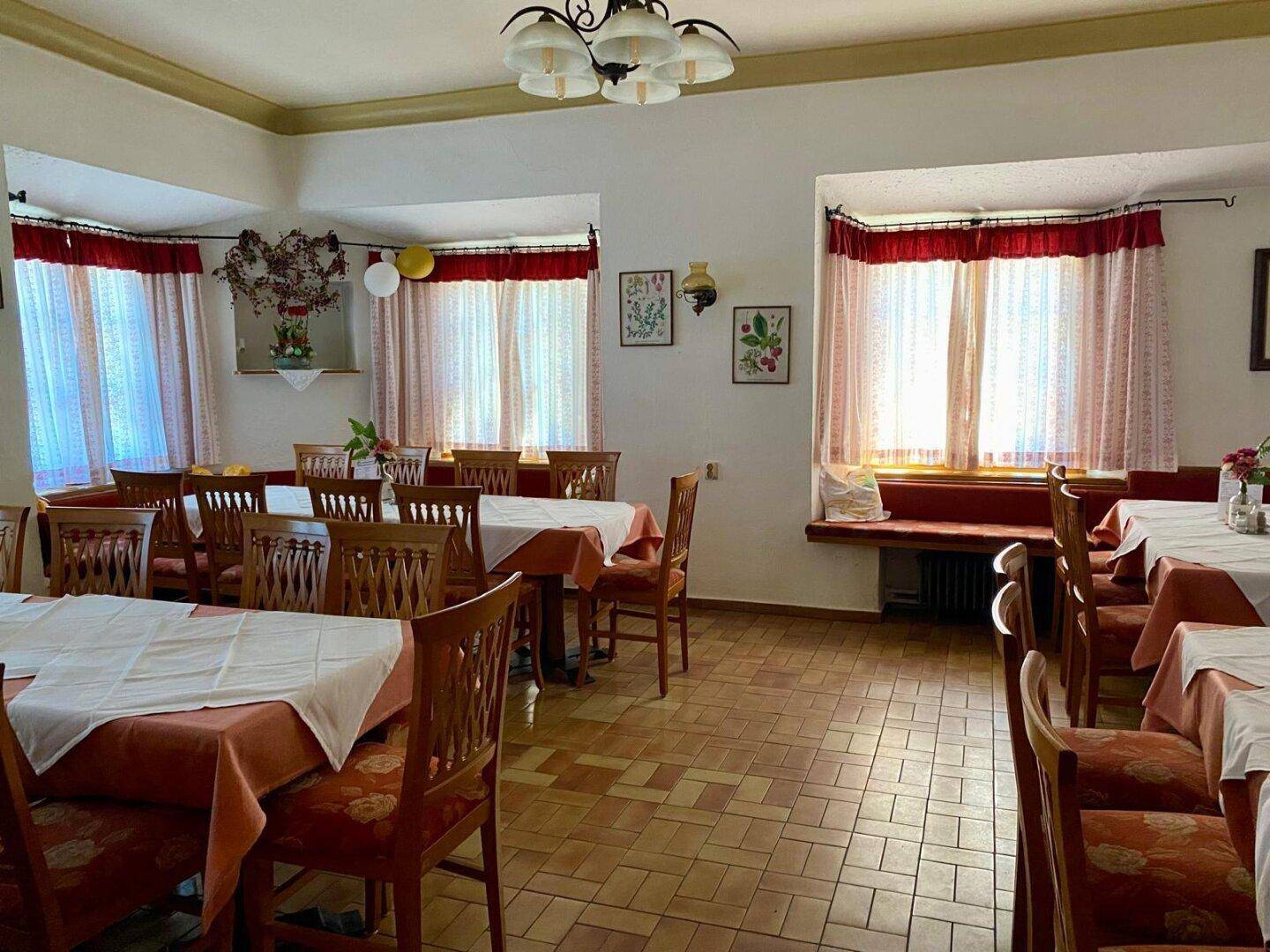 Heller Essbereich mit vielen Tischen, traditionellen Holzstühlen und Fenstern mit roten Vorhängen.