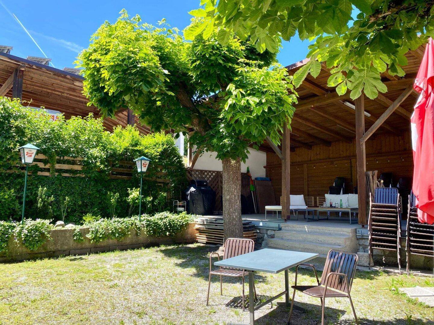 Gemütlicher Außenbereich mit einem schattenspendenden Baum, einem Tisch und Stühlen sowie einer überdachten Holzterrasse.