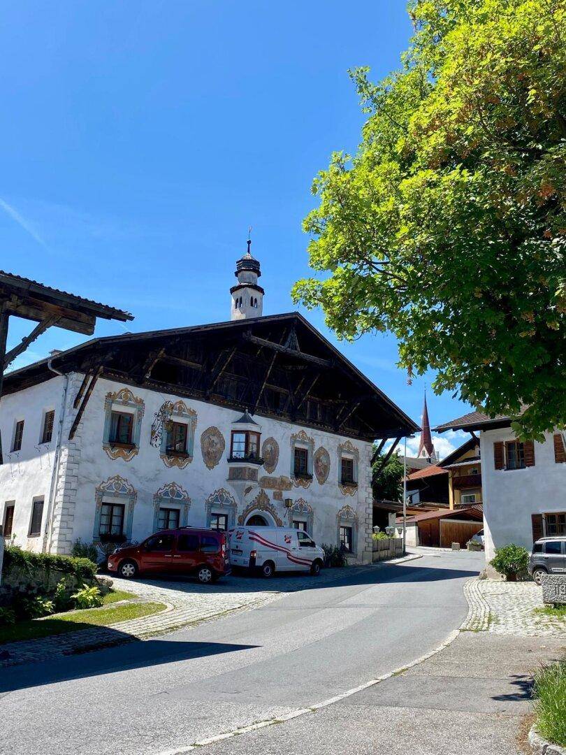 Historisches Gebäude mit bemalter Fassade und Holzbalkonen an einer gepflasterten Straße in einer ländlichen Umgebung.
