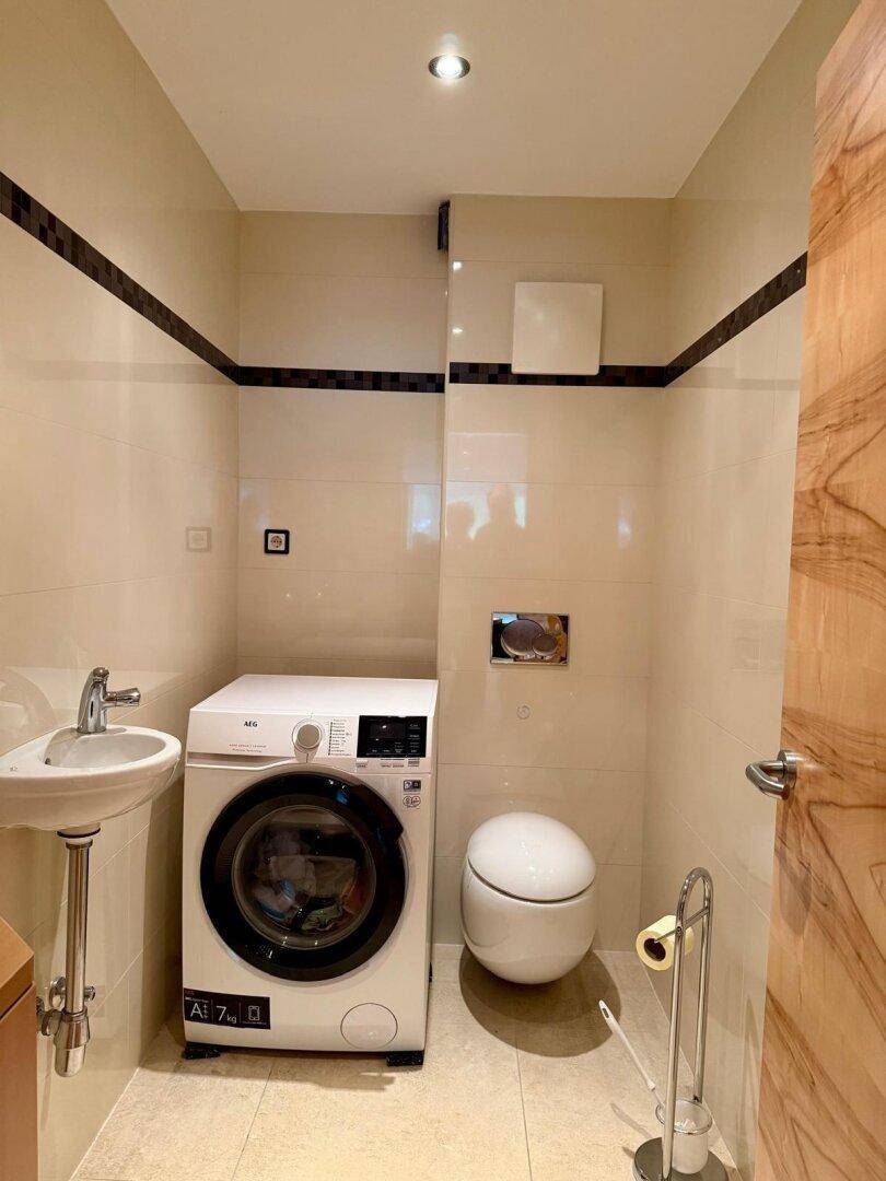 Kleines Gäste-WC mit Waschmaschine, wandhängendem WC und hellem Fliesenboden.