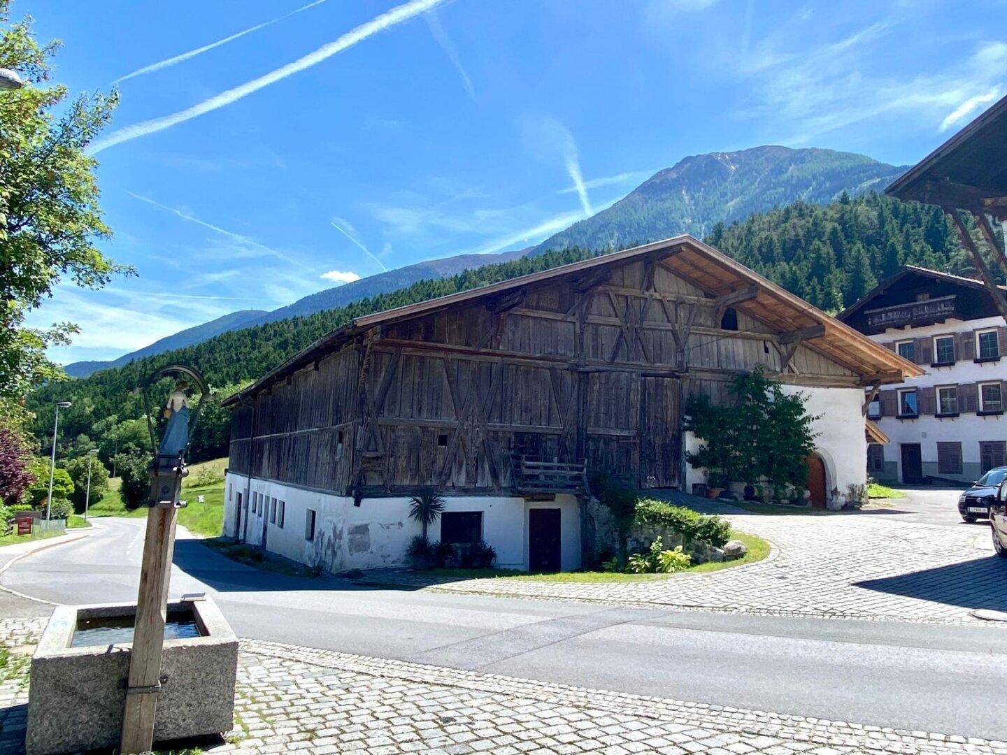Großes Holzgebäude mit traditioneller Bauweise und einem Brunnen im Vordergrund, umgeben von Bergen und Natur.
