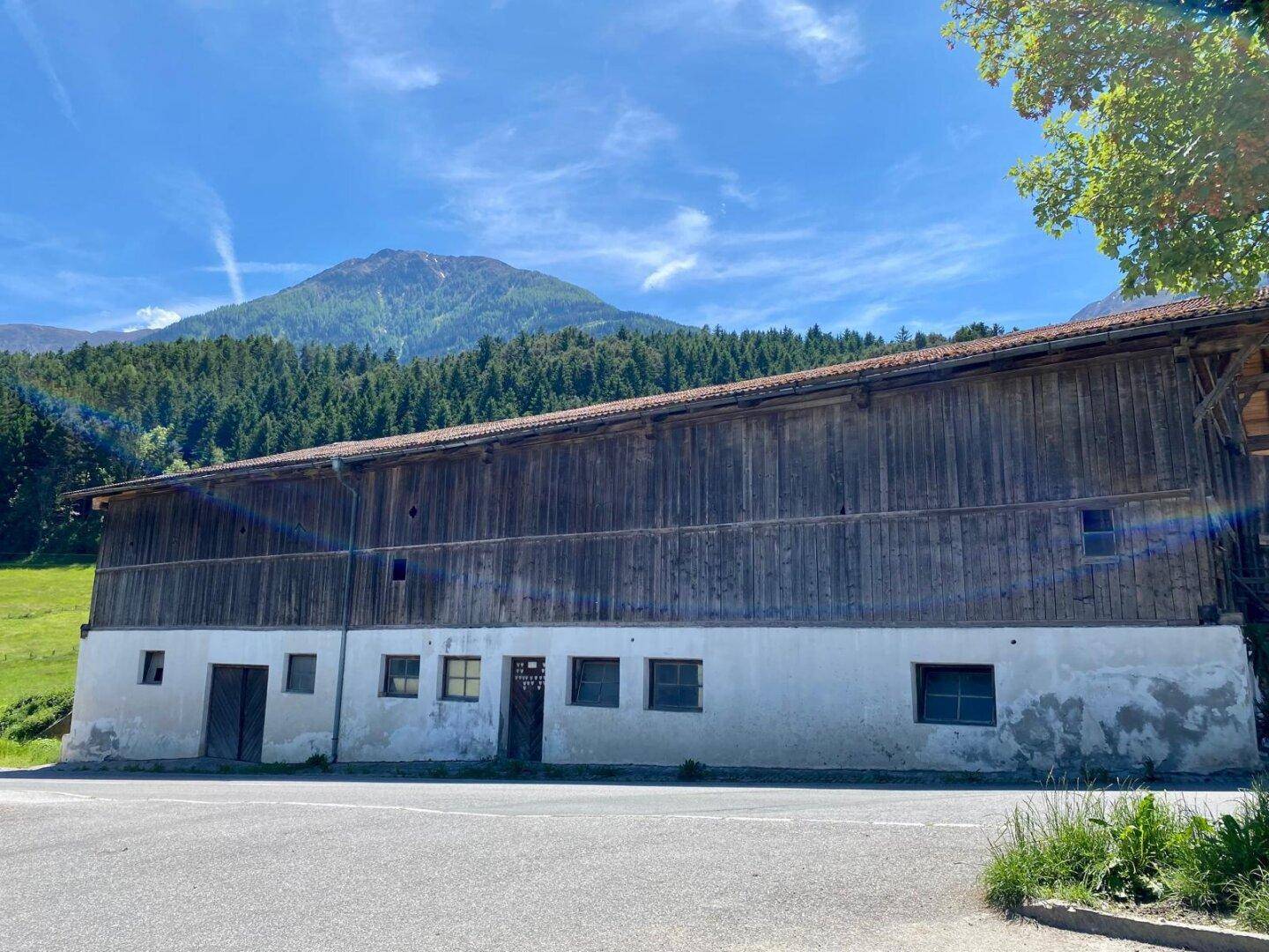 Lange Seite eines traditionellen Holzgebäudes mit weiß getünchtem Sockel und Blick auf grüne Hügel und Berge.