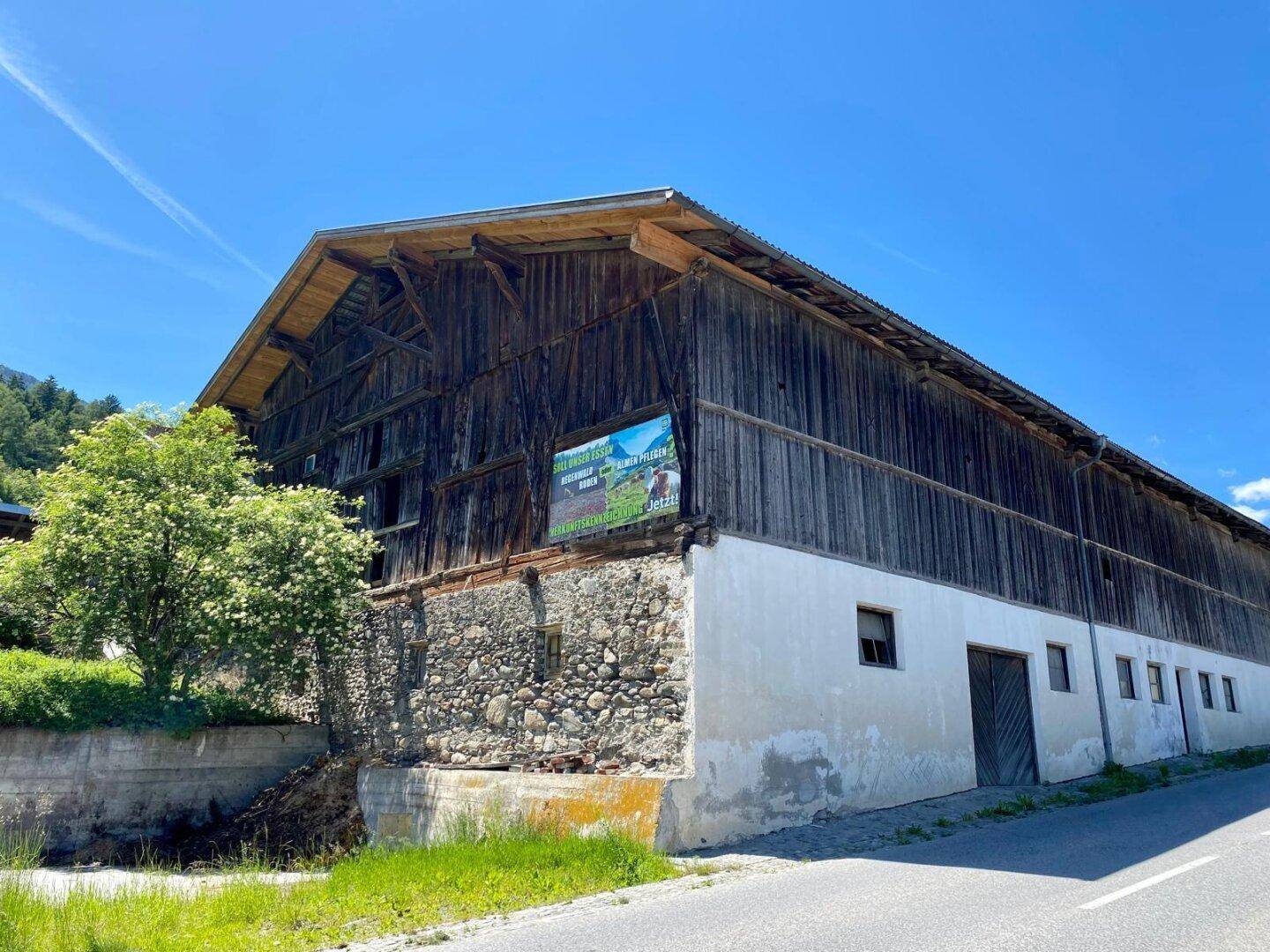 Rustikales Holzgebäude mit Steinfundament und einem Werbebanner an der Seite, umgeben von grüner Landschaft.