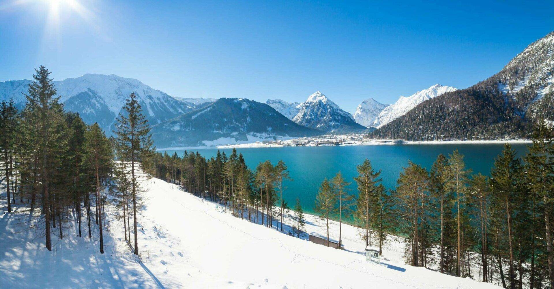 Winterliche Berglandschaft mit schneebedeckten Hängen, einem klaren See und majestätischen Gipfeln unter strahlendem Sonnenschein.