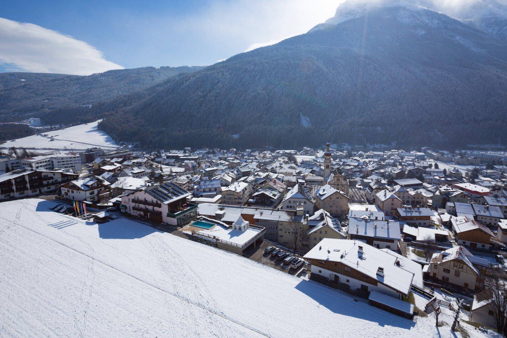 Luftaufnahme eines verschneiten Dorfes mit vielen Häusern und einem Berg im Hintergrund an einem sonnigen Wintertag.
