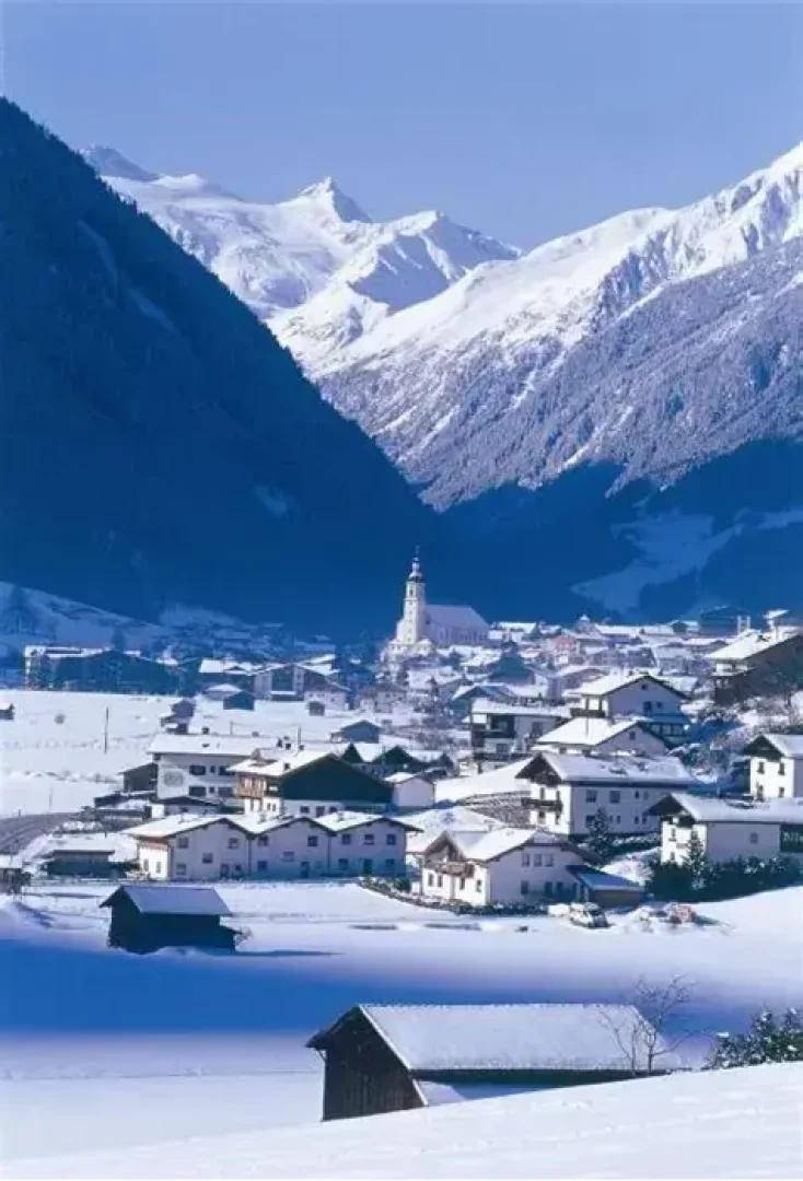 Winterlandschaft mit einem verschneiten Dorf in einem Tal, umgeben von majestätischen Bergen und einem Kirchturm.