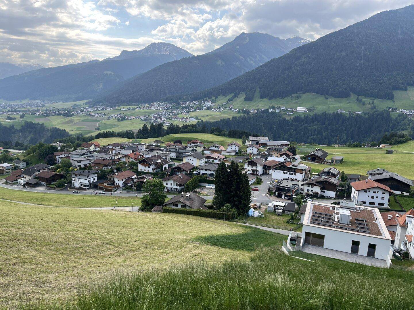 Eine weite Panoramaansicht eines Dorfes in einem Tal, umgeben von majestätischen Bergen und grünen Feldern.