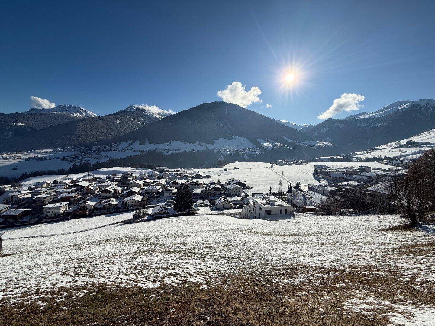 Eine winterliche Landschaft mit einem verschneiten Dorf in einem Tal, umgeben von schneebedeckten Bergen unter blauem Himmel.