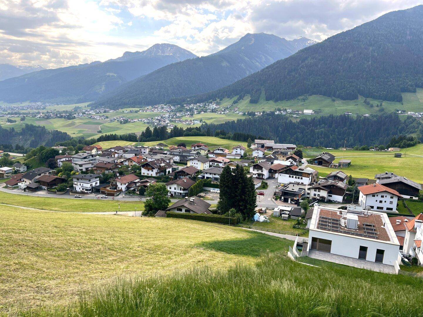 Eine weite Panoramaansicht eines Dorfes in einem Tal, umgeben von majestätischen Bergen und grünen Feldern.