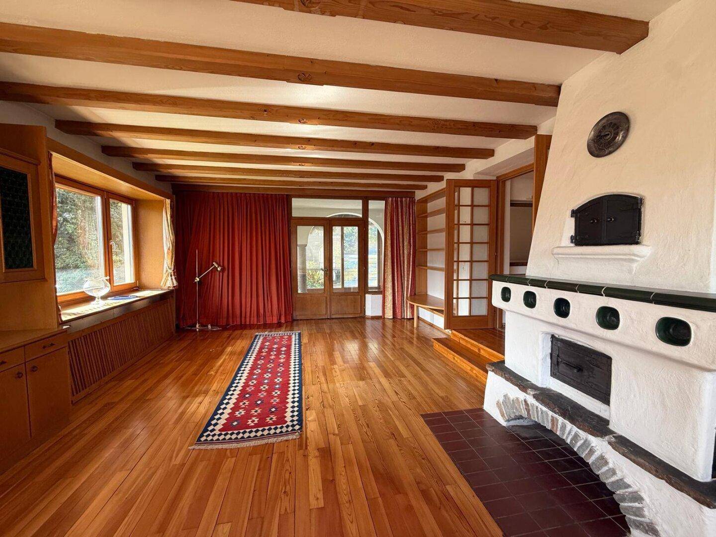 Geräumiges Wohnzimmer mit Holzbalken, Parkettboden und einem markanten Kachelofen als Mittelpunkt.