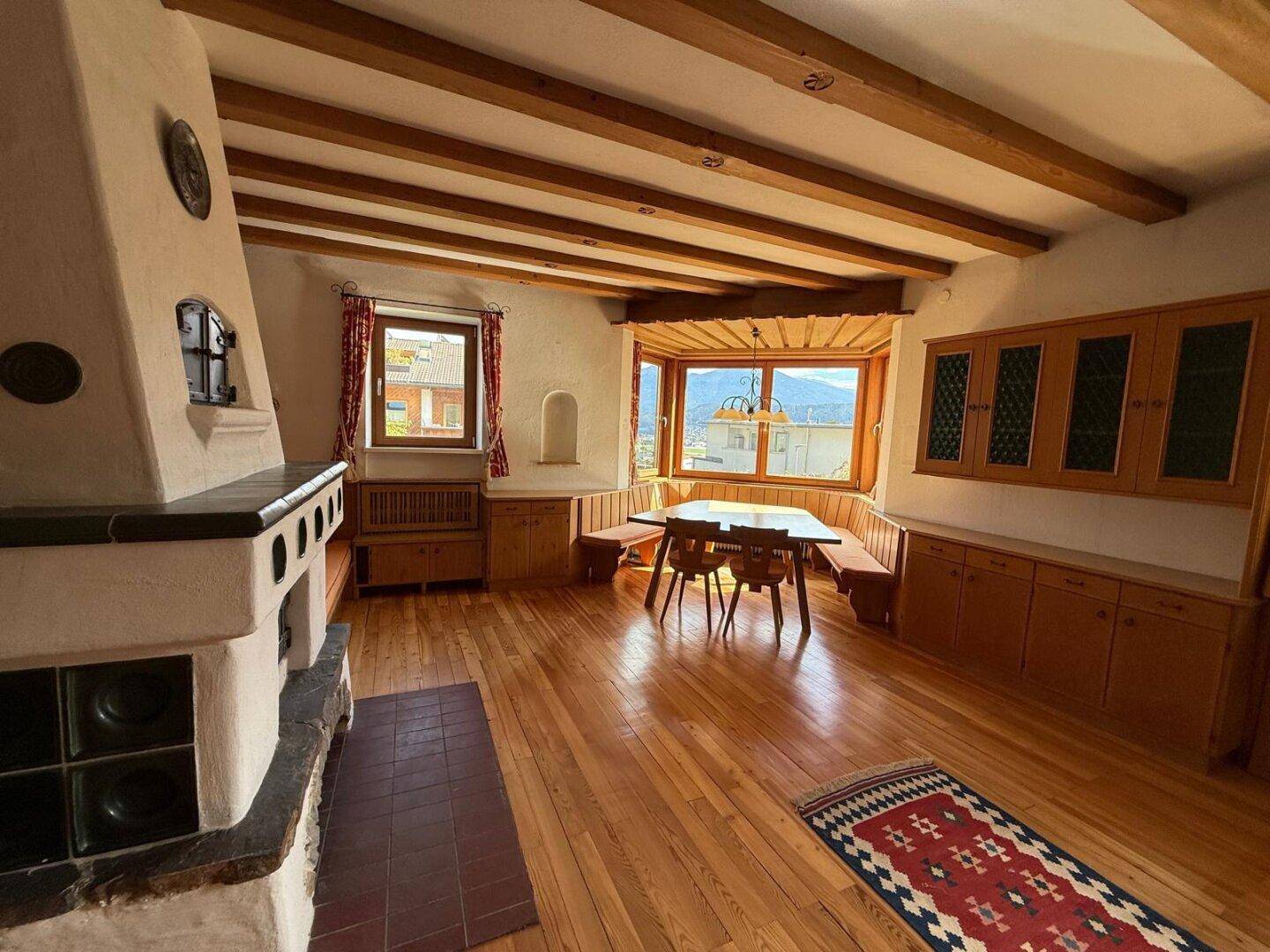 Traditioneller Wohn-Essbereich mit Holzbalkendecke, Kachelofen und Sitzbank am Fenster mit Aussicht.