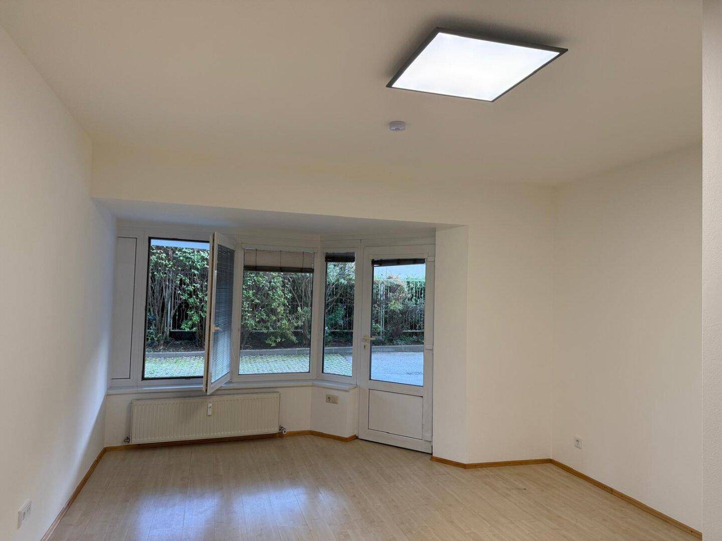 Heller Wohnraum mit Laminatboden und großer Fensterfront, die viel Tageslicht hereinlässt und Zugang nach draußen bietet.