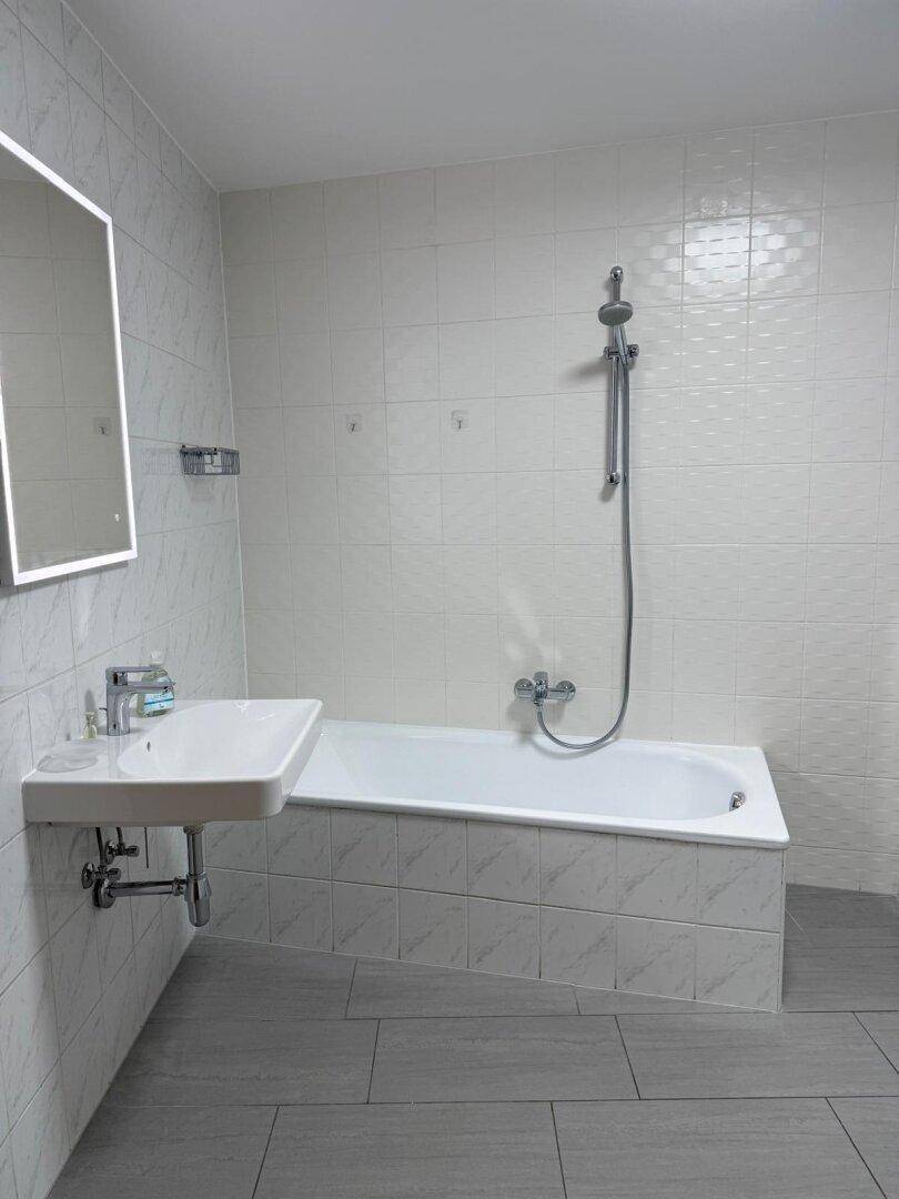 Helles Badezimmer mit Badewanne, Duscharmatur und modernem Waschbecken, umgeben von weißen Wandfliesen.