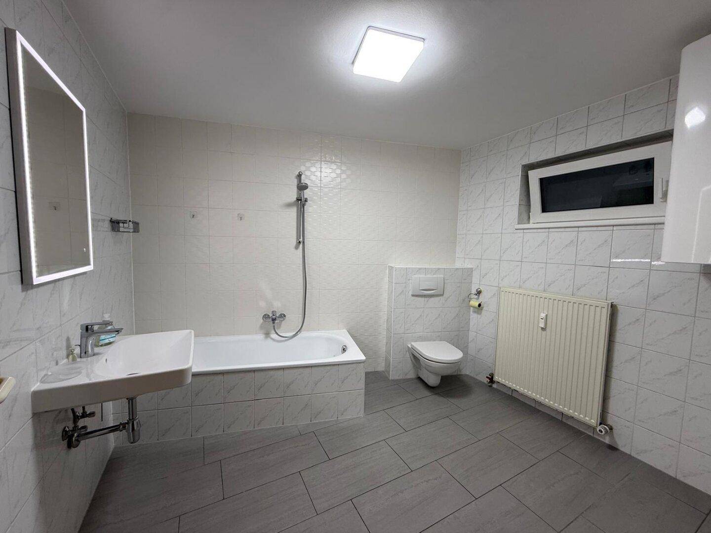 Modern gefliestes Badezimmer mit Badewanne, WC, Waschbecken und Fenster für natürliche Belüftung.