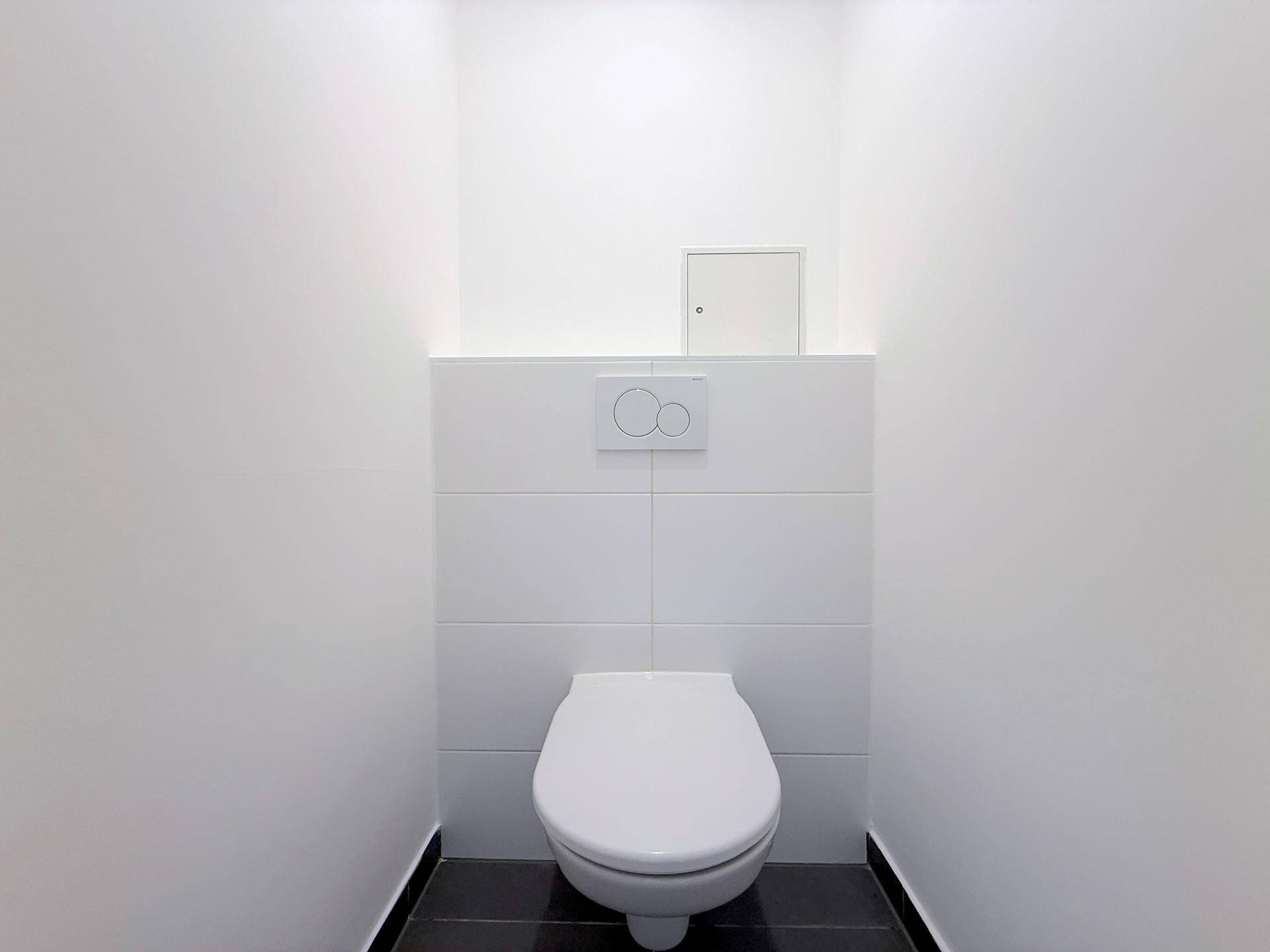 Separates WC mit wandmontierter Toilette und dunklen Bodenfliesen in einem schmalen Raum.