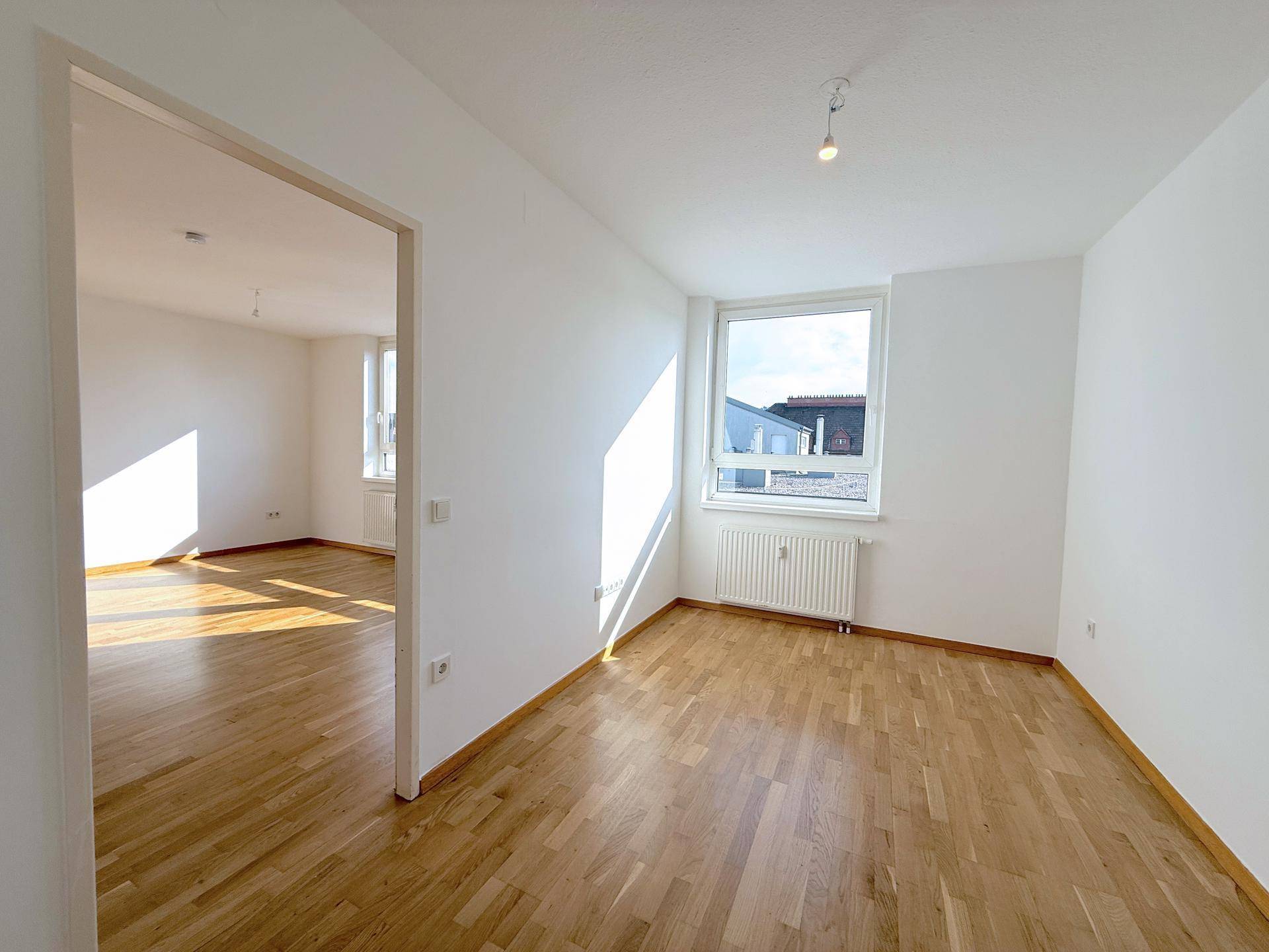 Leeres Zimmer mit Parkettboden und einem Fenster, das viel Sonnenlicht in den Raum bringt.