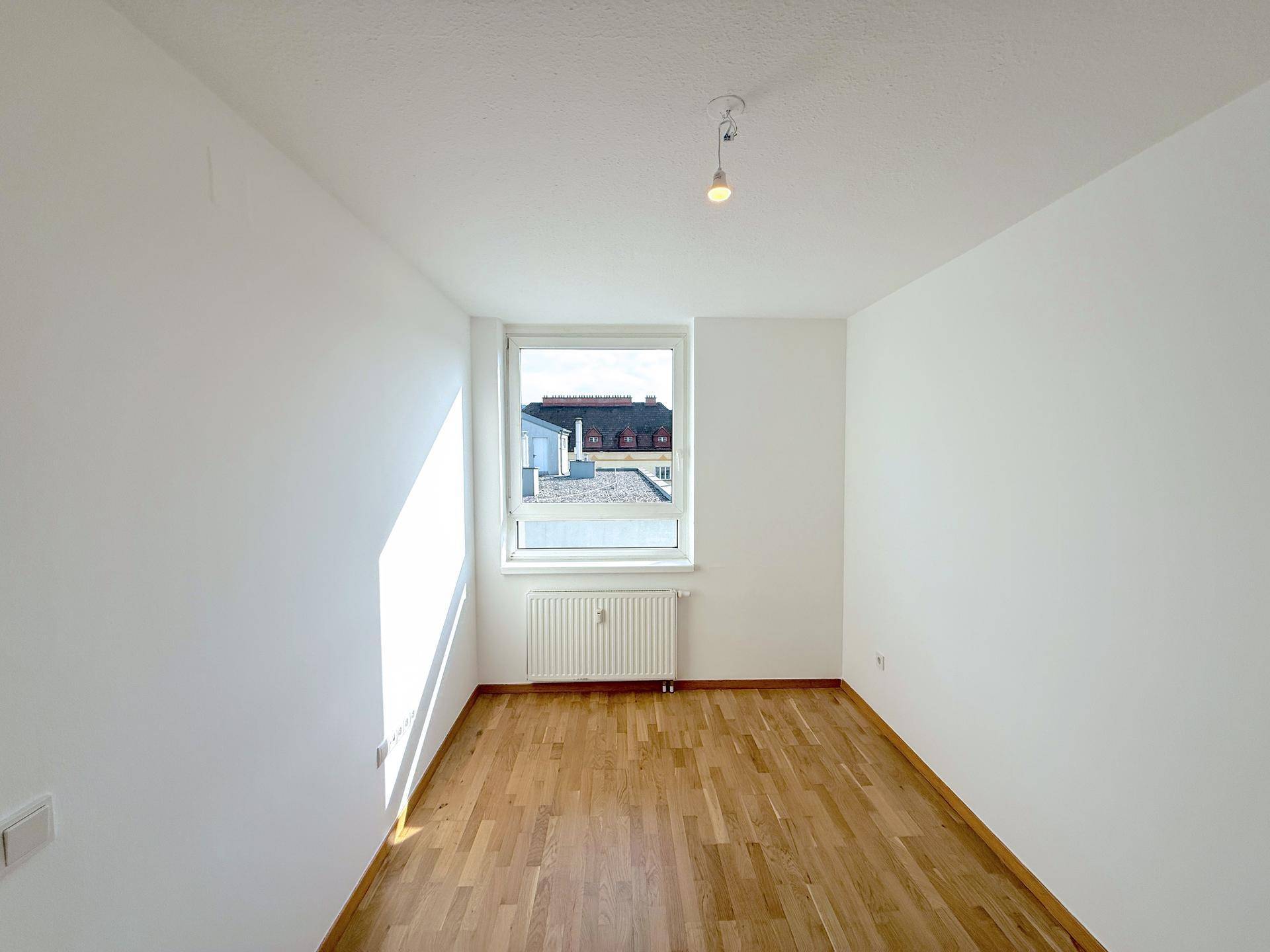 Helles, leeres Zimmer mit Parkettboden und einem Fenster, das viel Tageslicht hereinlässt.