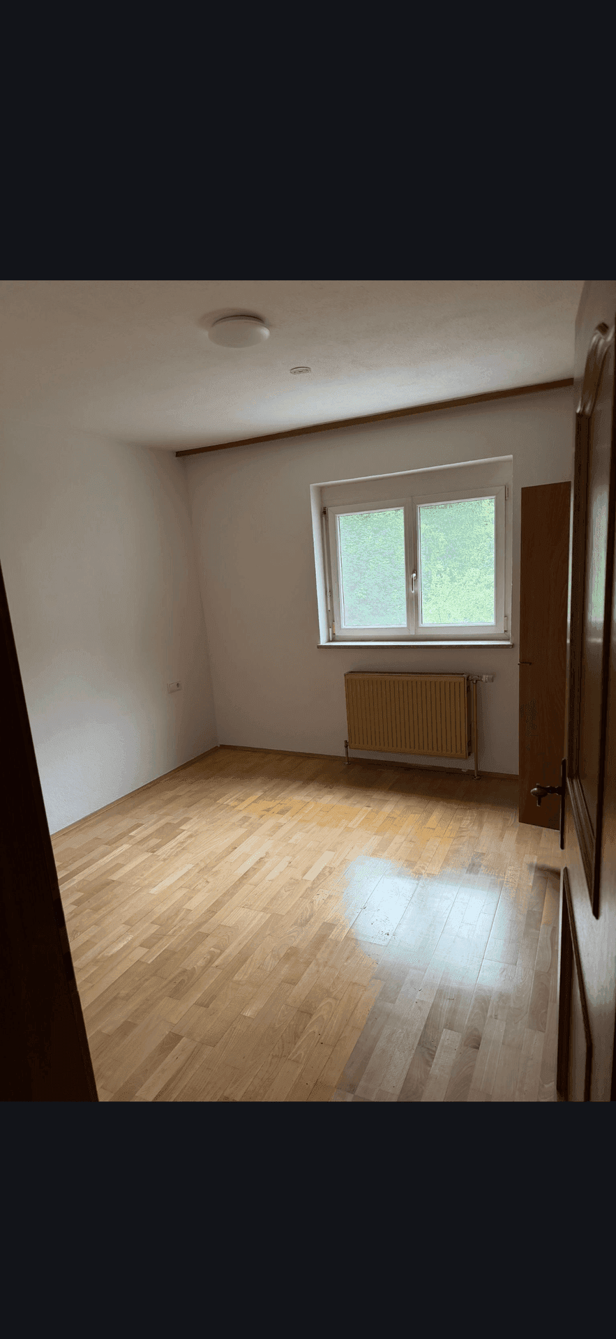 Leeres Zimmer mit hellem Holzboden, Fenster und Heizkörper, ideal als Schlaf- oder Arbeitszimmer.