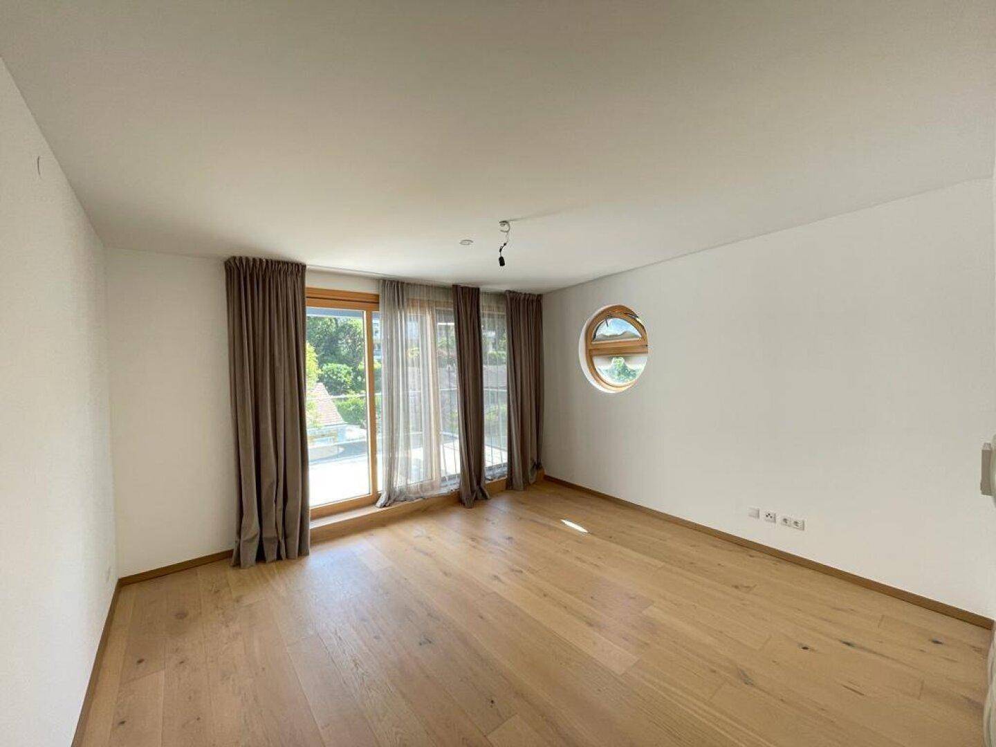 Helles Zimmer mit Holzboden, großen Fenstern und einem runden Akzentfenster.