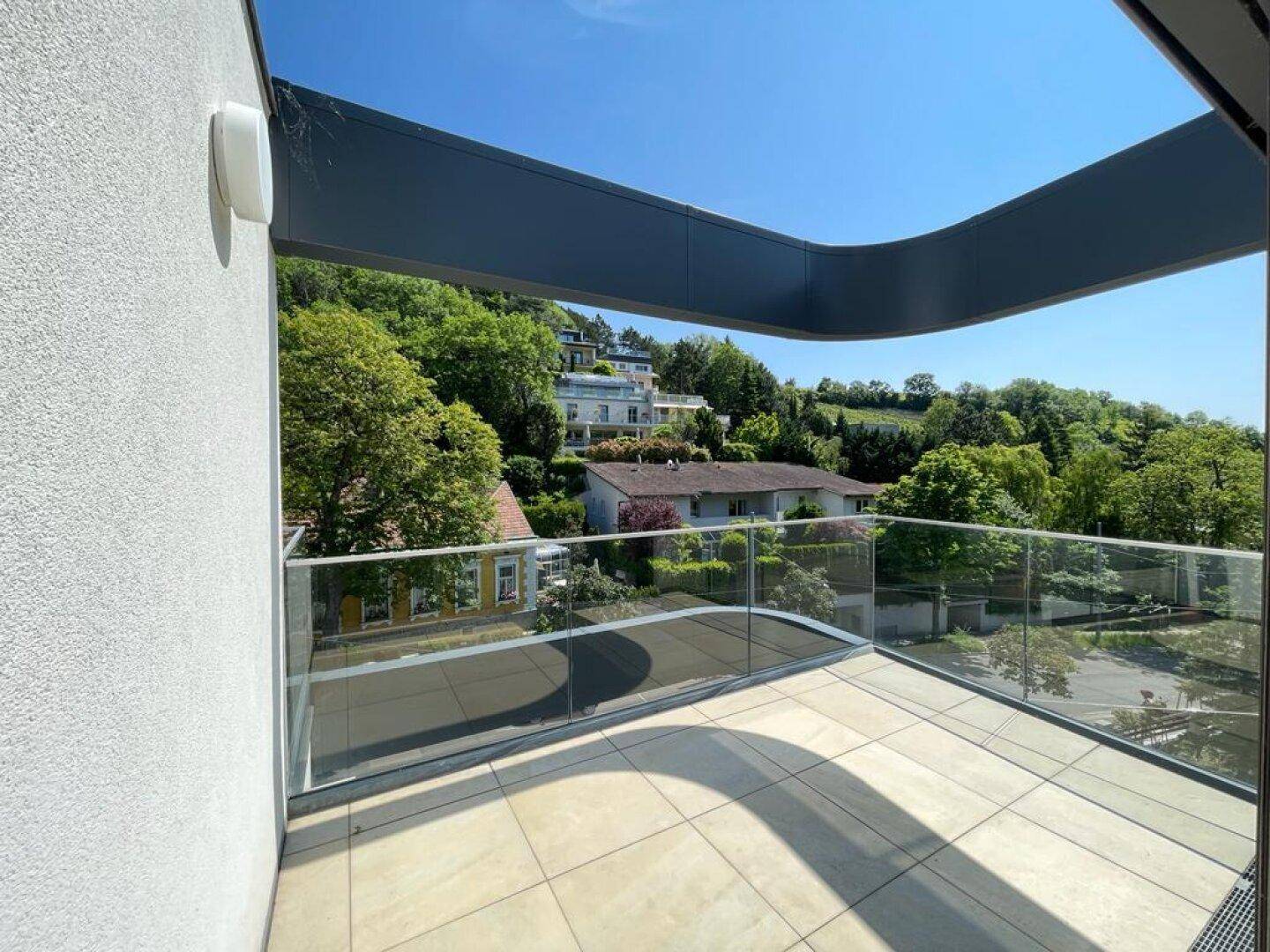 Heller Balkon mit Glasgeländer und weitem Blick auf grüne Hügel und umliegende Häuser.