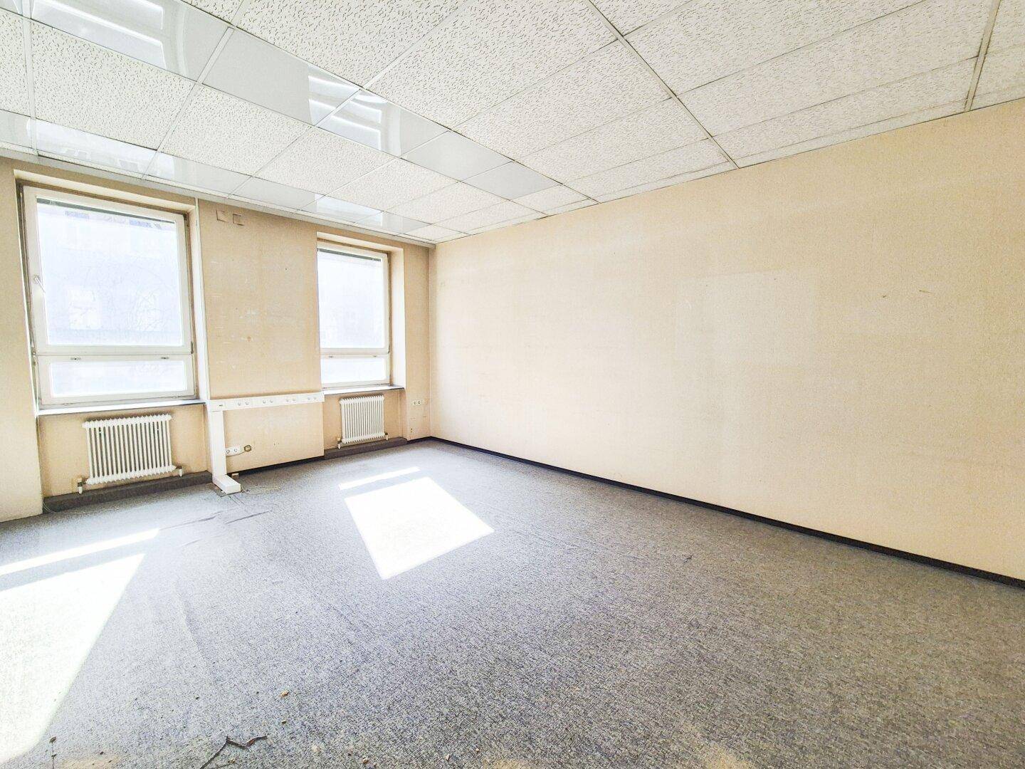 Leeres Büro mit Teppichboden, beigen Wänden und zwei Fenstern mit Heizkörpern darunter.