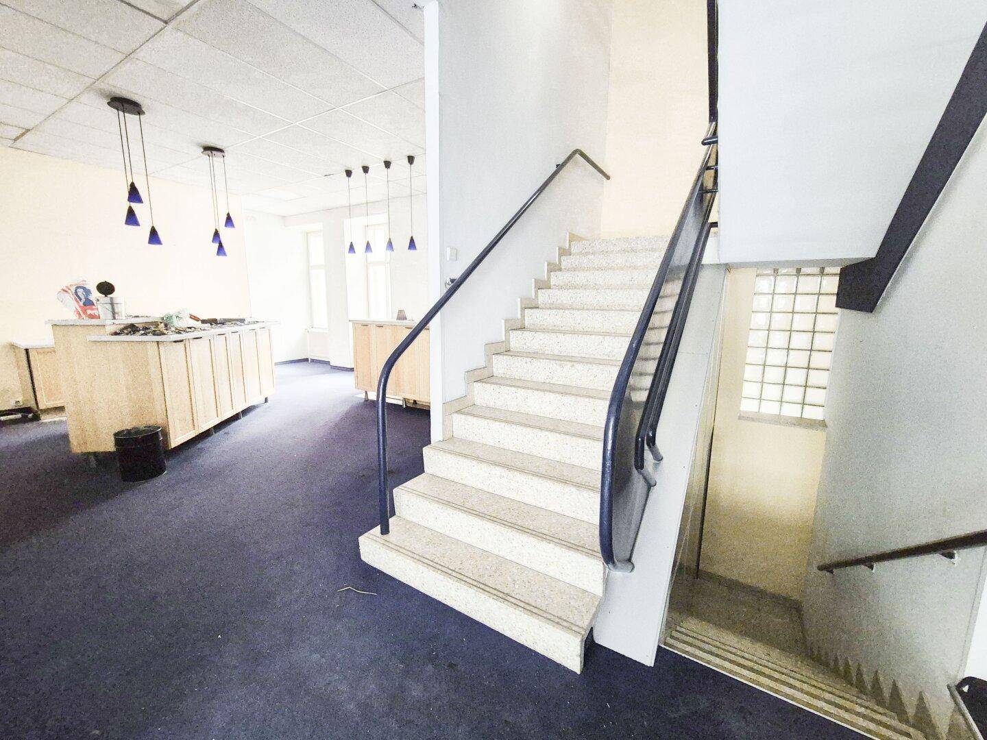 Treppenhaus mit Teppichboden und einer Theke im Hintergrund, beleuchtet von blauen Pendelleuchten.