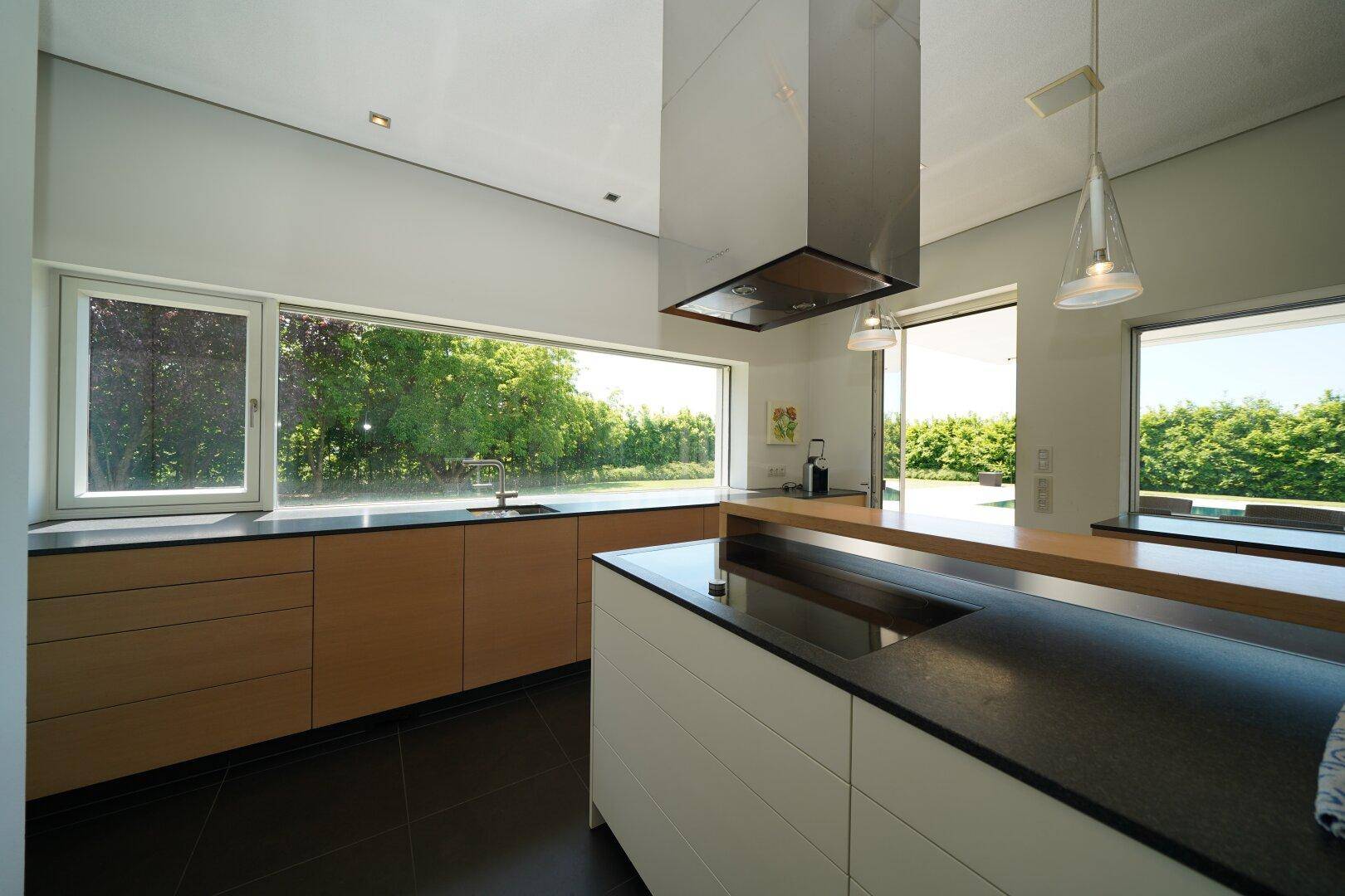 Moderne Küche mit Holzfronten, dunklen Arbeitsplatten und weitem Blick ins Grüne durch große Fenster.