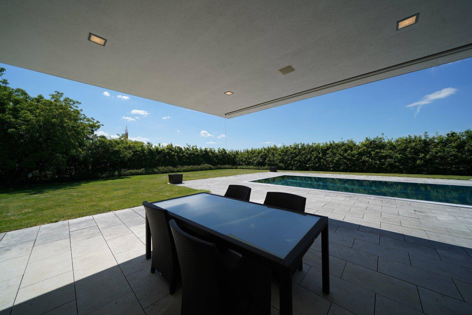 Überdachte Terrasse mit Esstisch, angrenzendem Rasen und Swimmingpool, umgeben von einer Hecke.