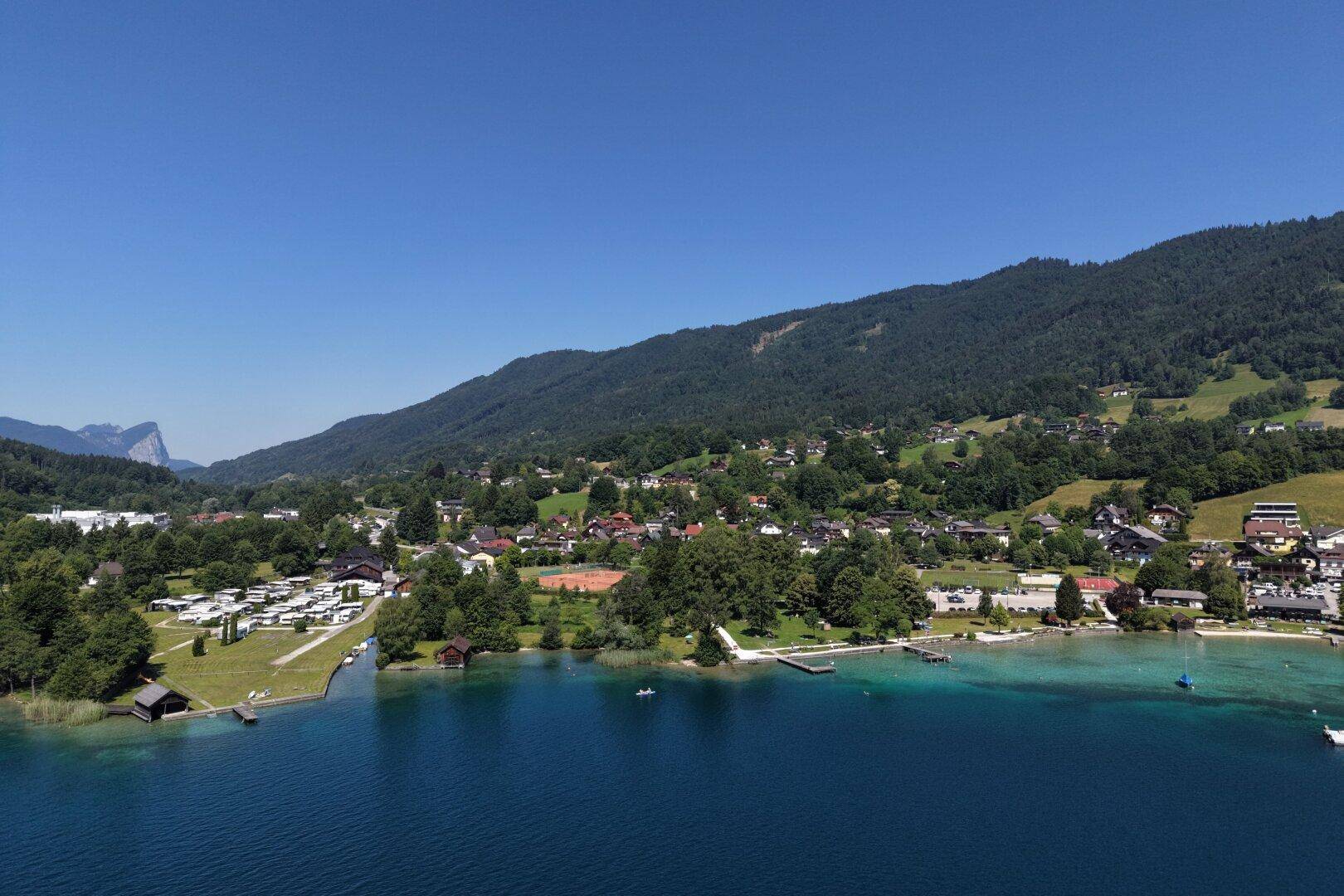 Weitläufiger Blick auf den See, die bewaldeten Berge und die Küstenbebauung unter blauem Himmel.