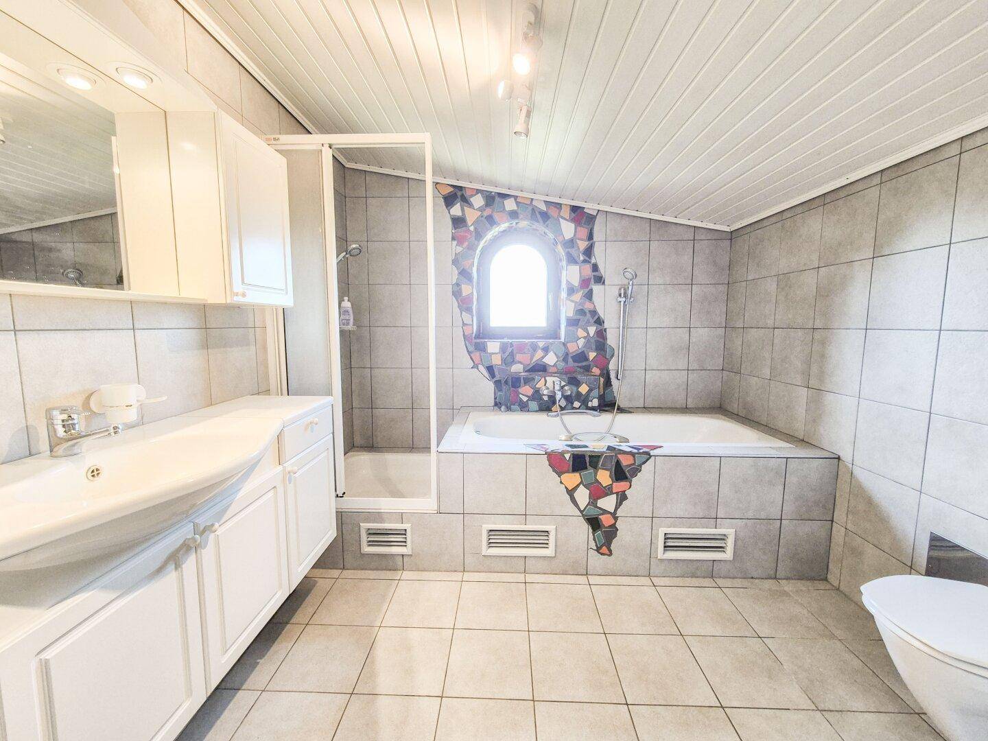 Geräumiges Badezimmer mit Badewanne, Dusche, Einzelwaschbecken und farbenfrohem Mosaikfenster.