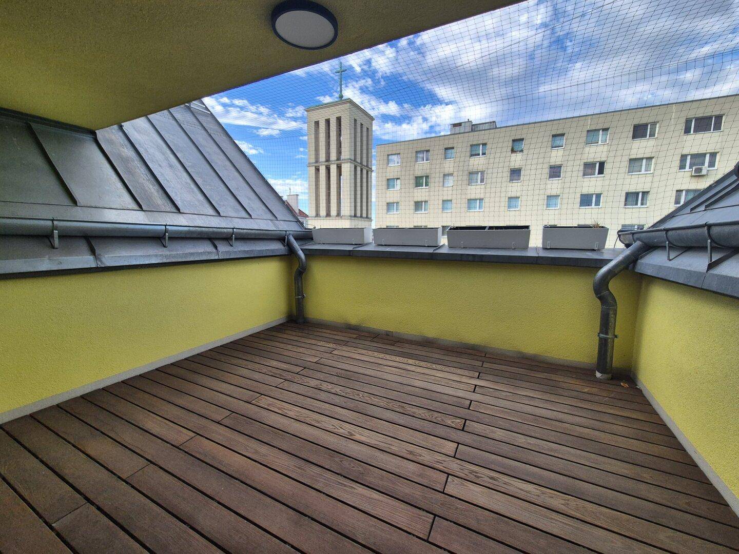 Kleiner Balkon mit Holzboden und Blick auf die umliegenden Gebäude und einen Kirchturm.