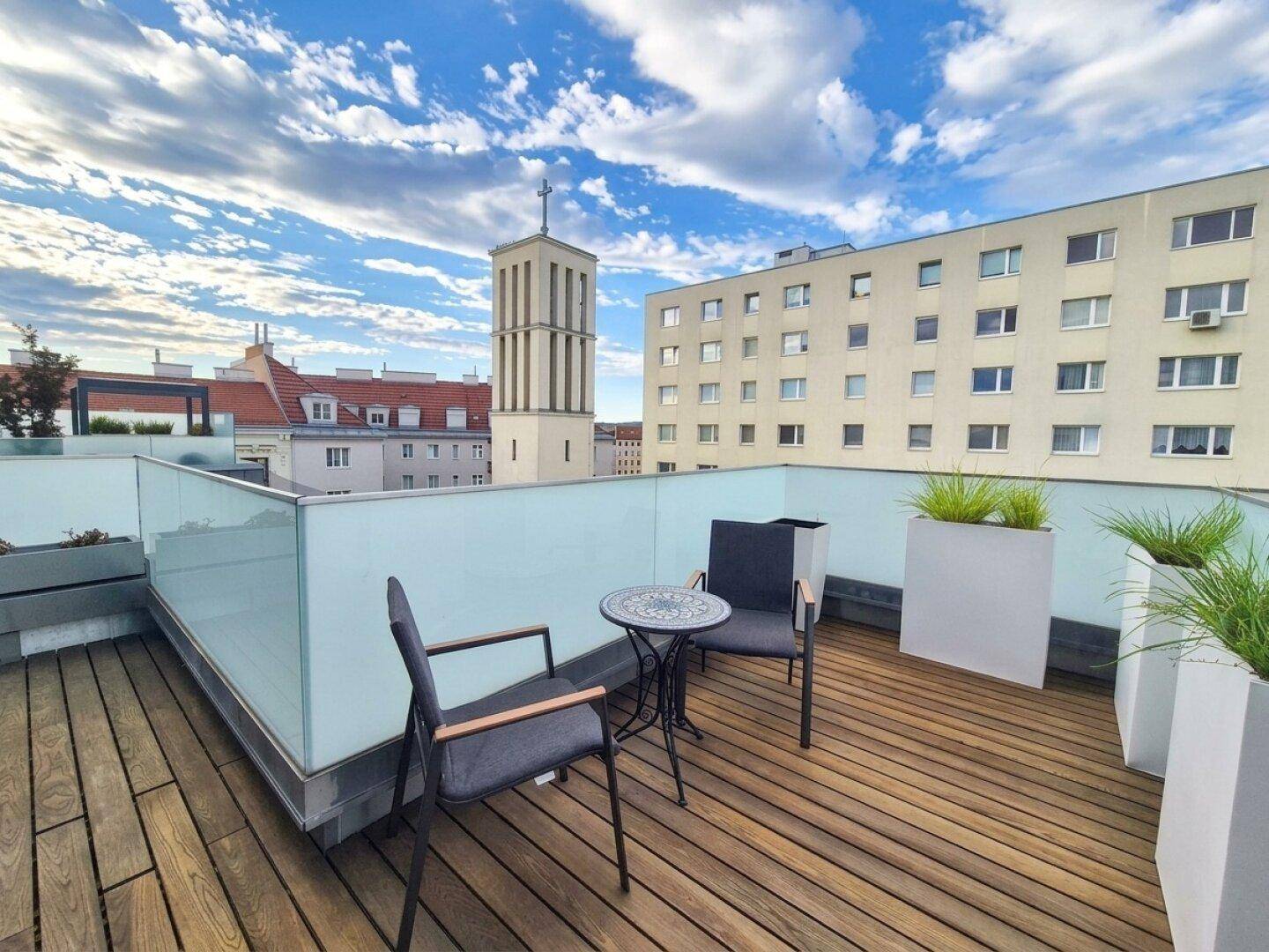Möblierte Dachterrasse mit Holzboden, Glasgeländer und weitem Blick über die Stadt.