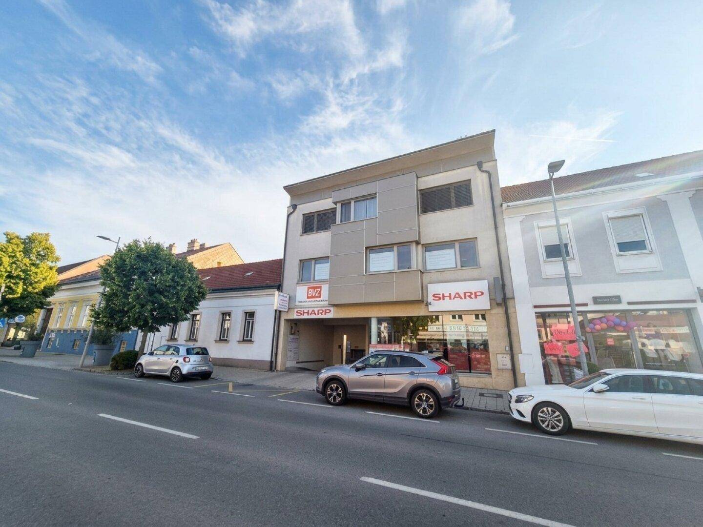 Gebäudeansicht von der Straße mit Geschäften im Erdgeschoss und parkenden Autos.