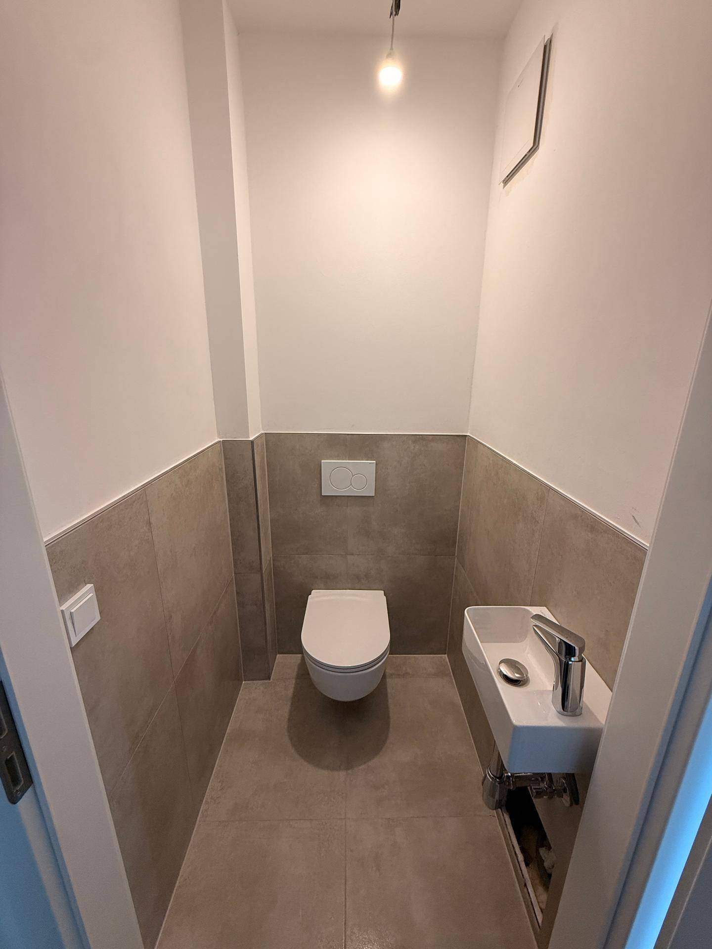 Modernes Gäste-WC mit Wand-WC, kleinem Waschbecken und grauen Fliesen an Wänden und Boden.