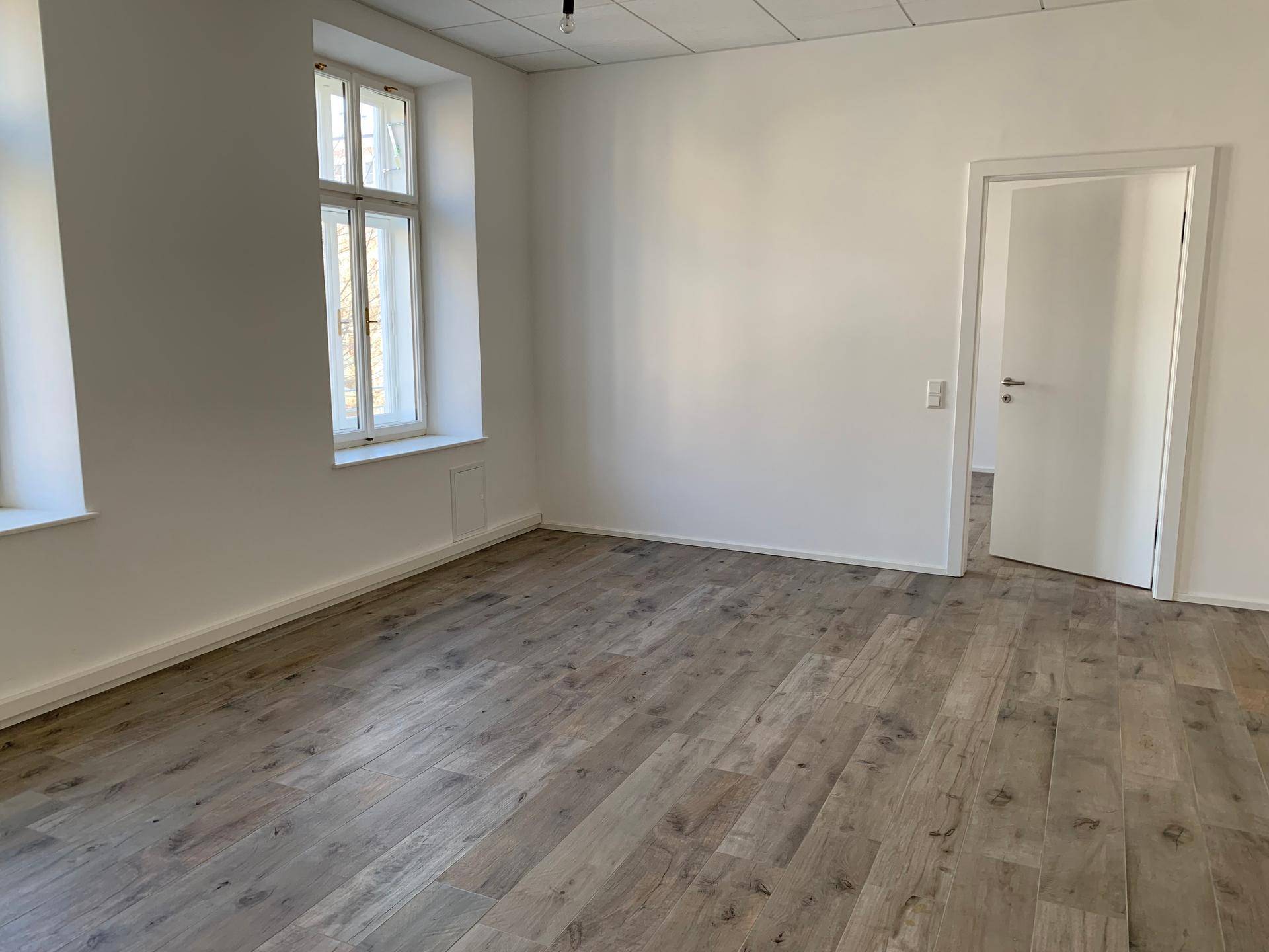 Heller, leerer Raum mit einem Fenster und Holzboden, der vielseitig genutzt werden kann.